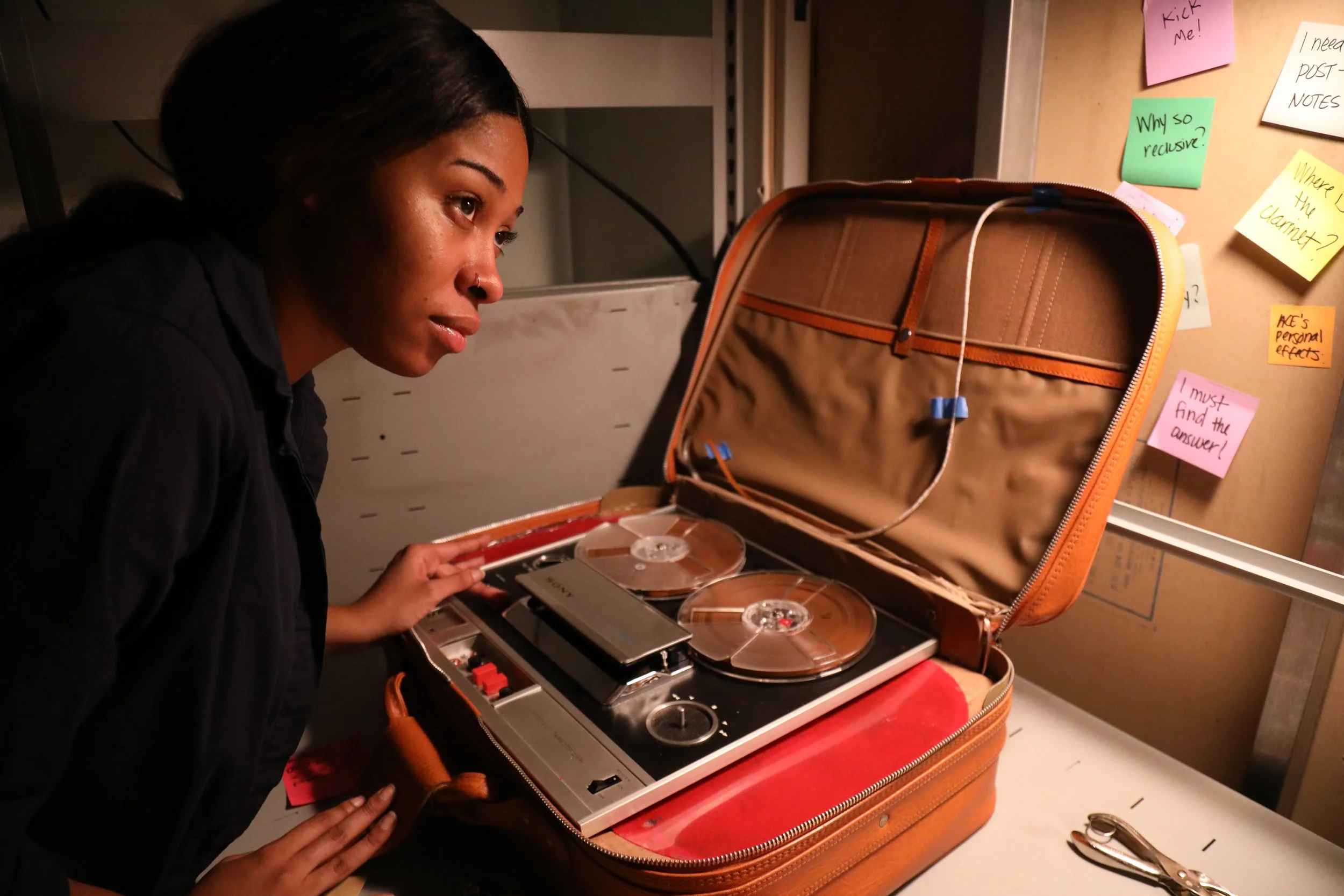 A woman with dark hair looks at a vintage film editing and music mixing device inside an orange briefcase on a table. The briefcase is open, revealing reels and controls, and is placed in front of a bulletin board with colorful sticky notes.
