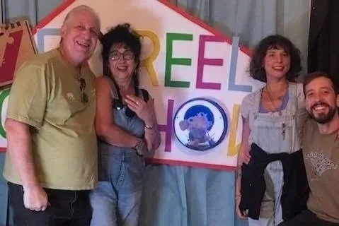 Group of four people standing in front of a colorful sign that says 'Reel Show' with a picture of a sheep. They are smiling and posing happily.
