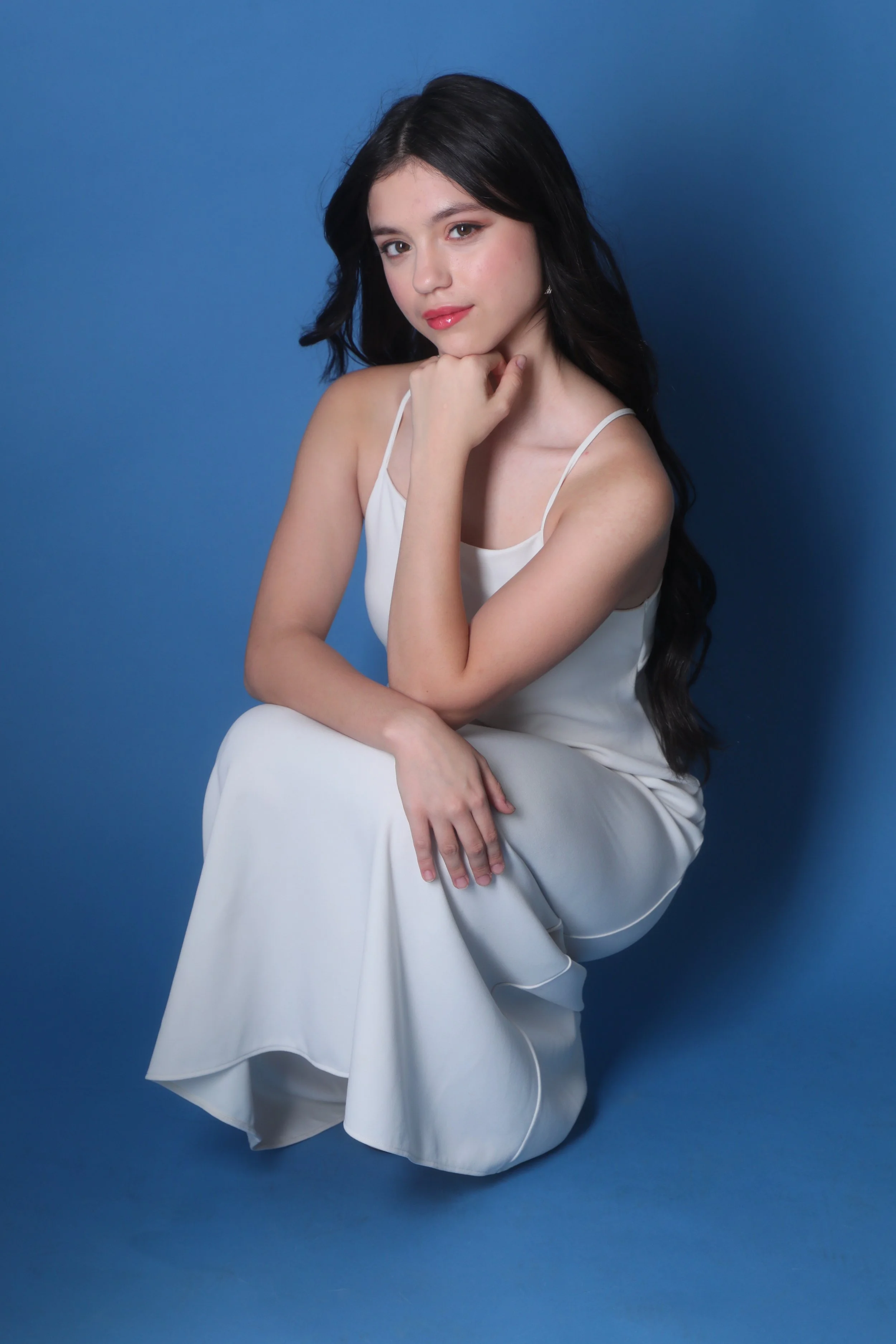 A young woman with long dark hair, sitting against a blue background, wearing a white dress with spaghetti straps, with her chin resting on her hand and a slight smile.