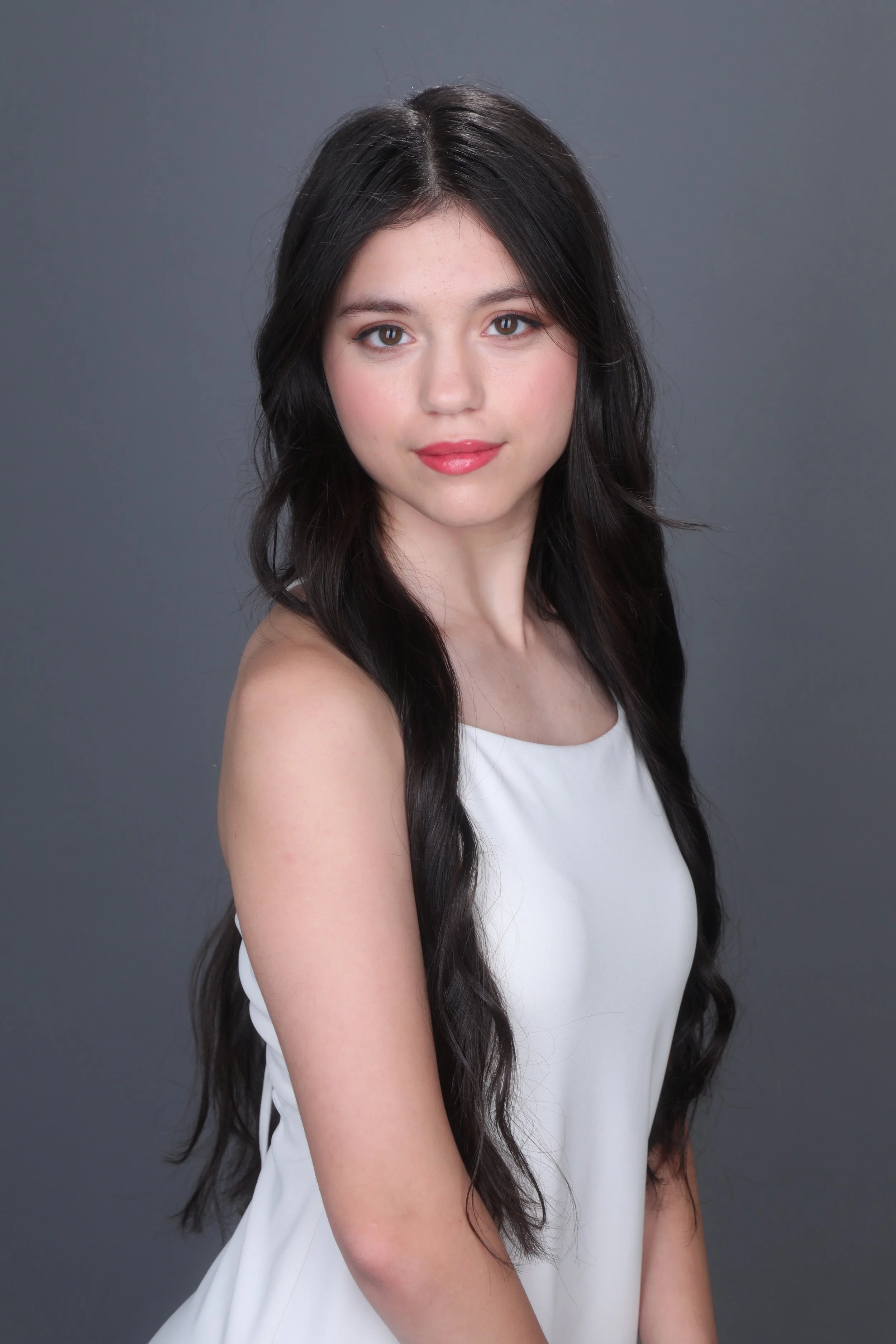 Portrait of a young woman with long dark wavy hair, wearing a white sleeveless top, smiling softly against a plain gray background.