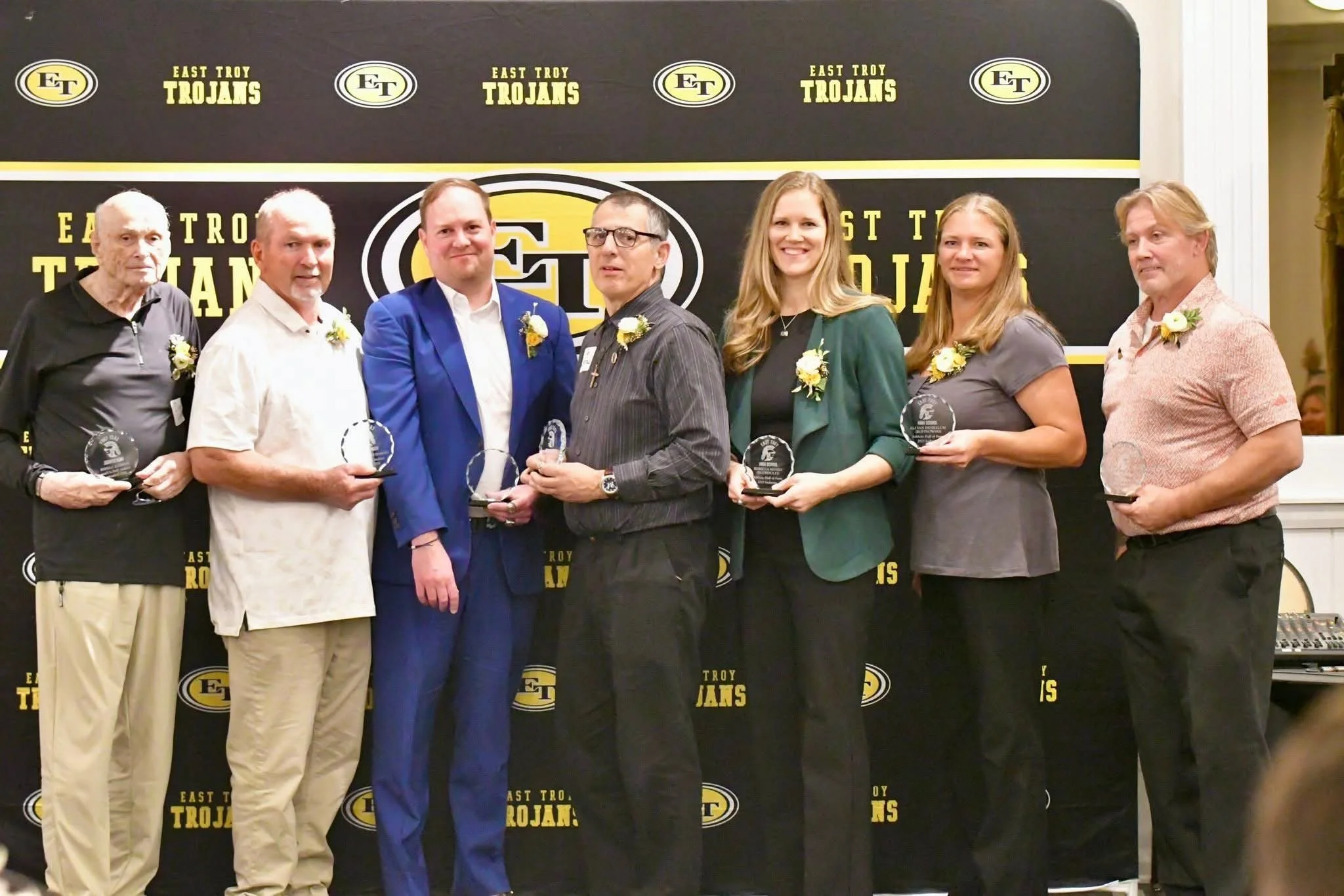 Group of seven people standing on stage at an awards ceremony, each holding a glass award, with a backdrop featuring 'East Troy Trojans' logo and text.