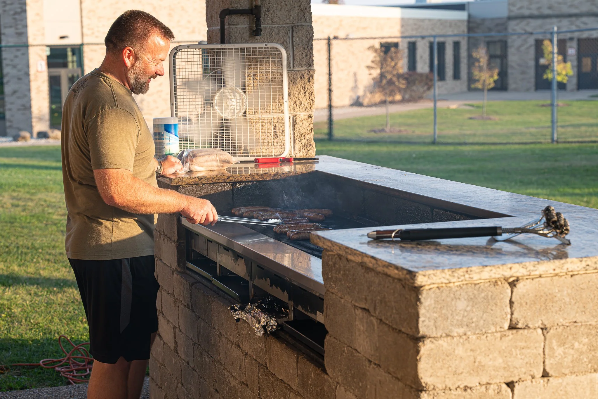 A man grilling sausages on an outdoor barbecue grill in a park or backyard during the daytime.