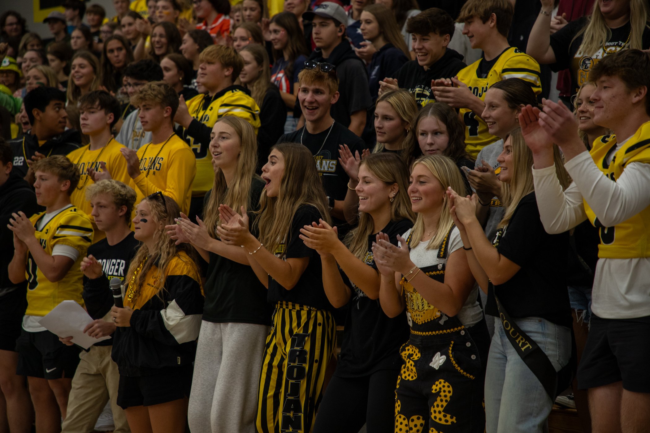 A group of excited and happy young people, many wearing yellow and black sports jerseys or team apparel, gathered together at a sports event, clapping and cheering.