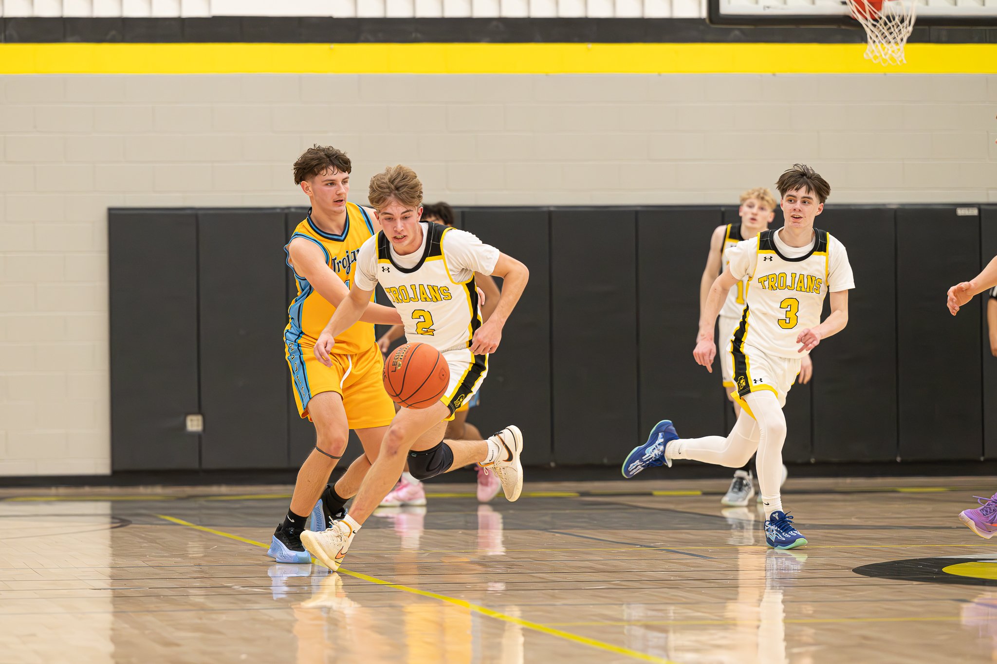 Young basketball players in a game, with two players wearing white jerseys labeled 'Trojans' competing for the ball against a player in a yellow and blue uniform, on an indoor court.