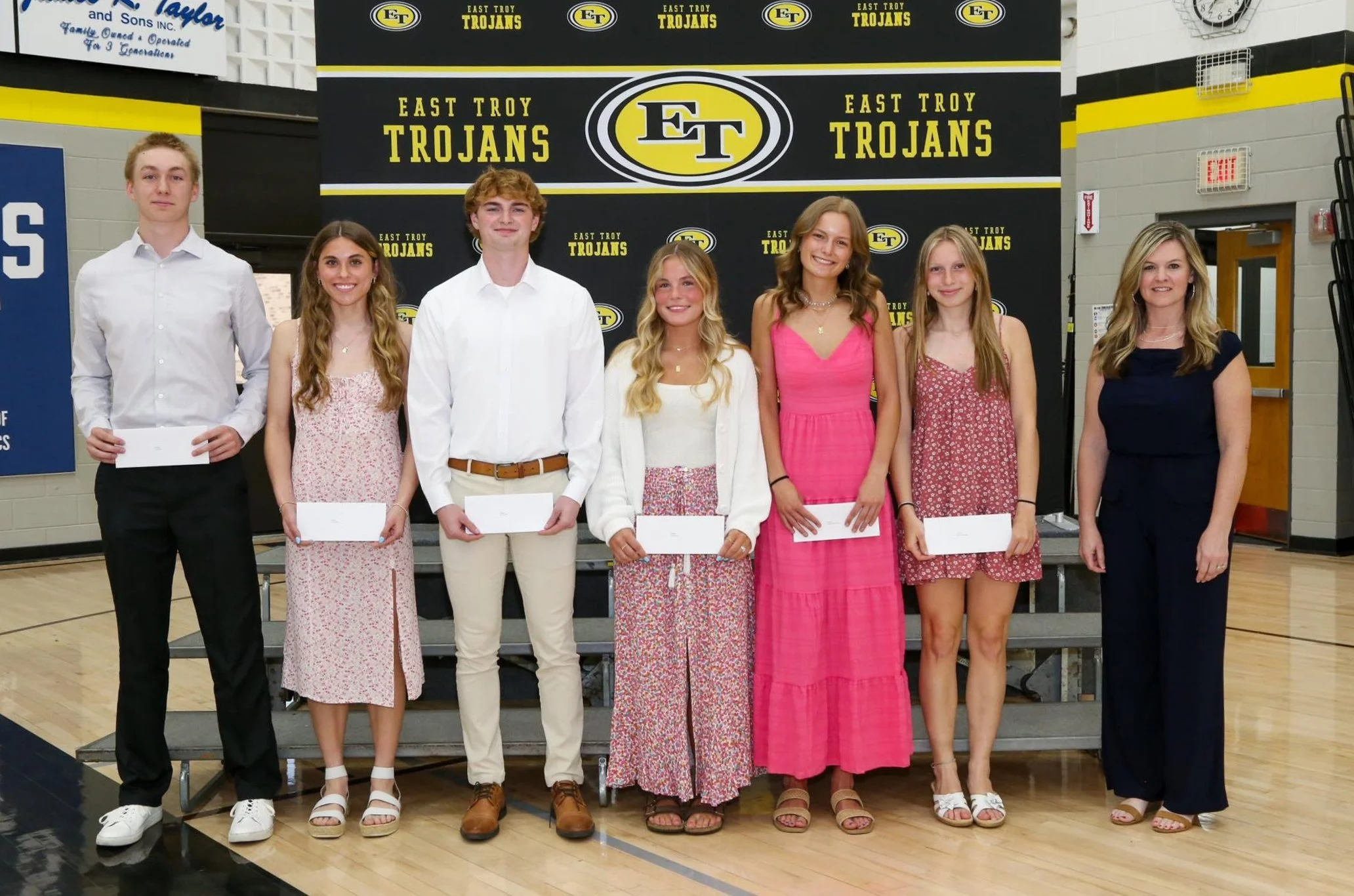 Group of seven students and one adult standing on a gymnasium stage for a photo, holding envelopes, with a background displaying 'East Troy Trojans' banners.