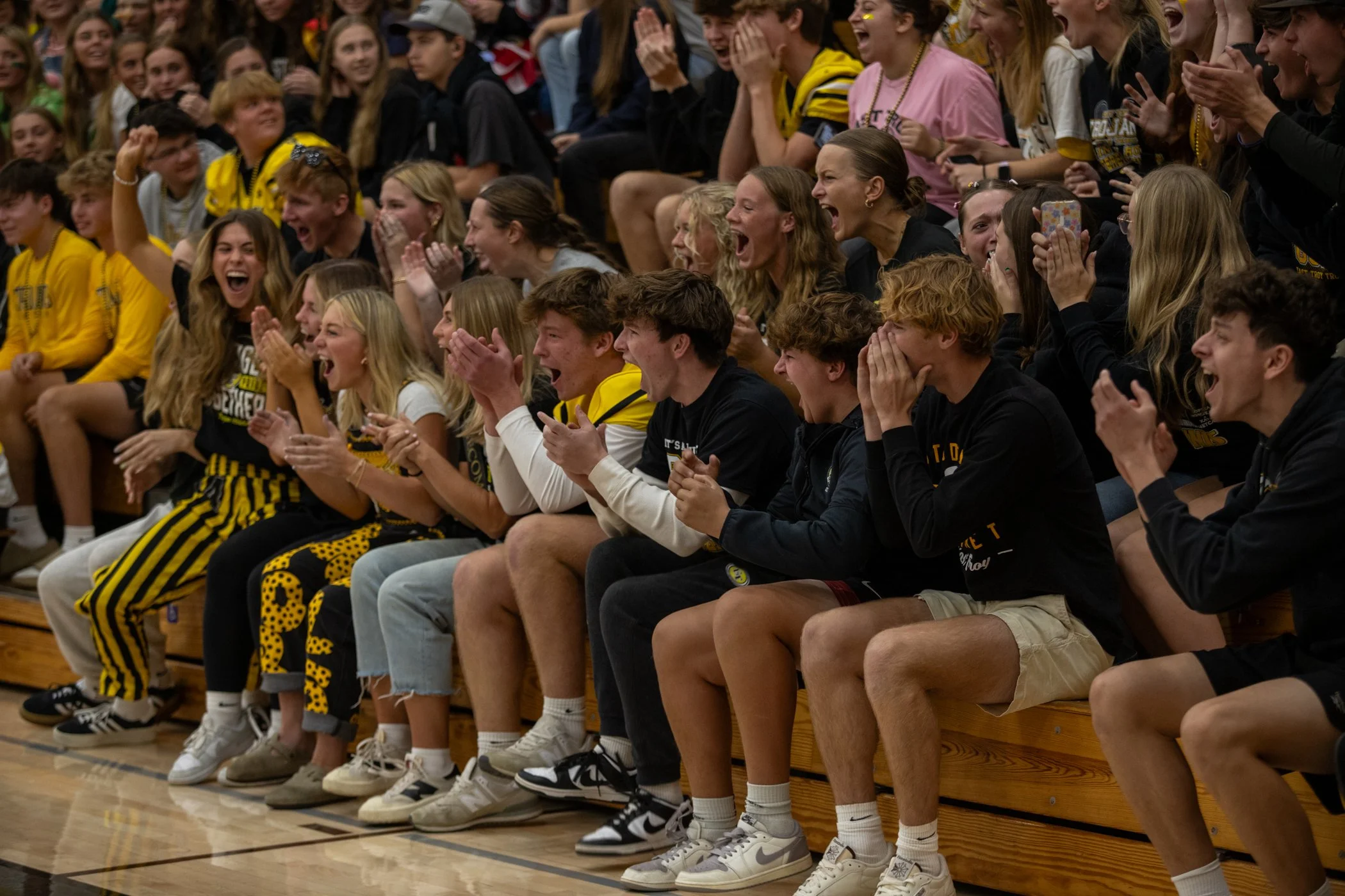Group of young people sitting on bleachers at a sporting event, laughing, cheering, and clapping, with some wearing black and yellow team apparel.
