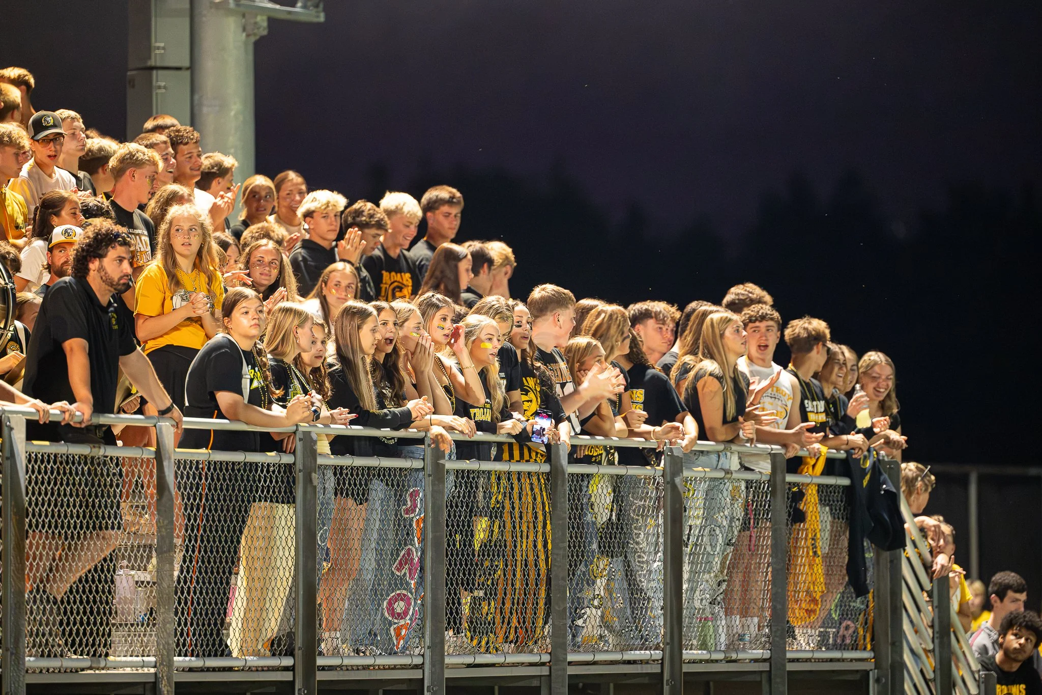 A group of high school students, wearing black and yellow clothing, standing along a chain-link fence at night, watching a sporting event with some cheering and taking photos.