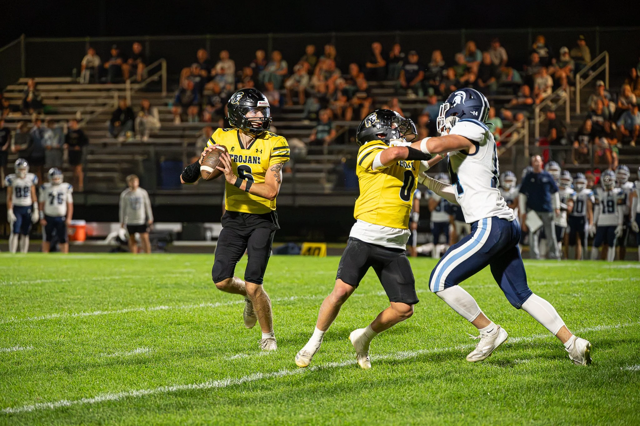 A football game at night with players on the field. A player in a yellow jersey holding a football looks to his left, while another player in yellow appears to be blocking a player in a white jersey. The player in white is attempting to block or tackle the player with the football. There are spectators sitting in the bleachers in the background.
