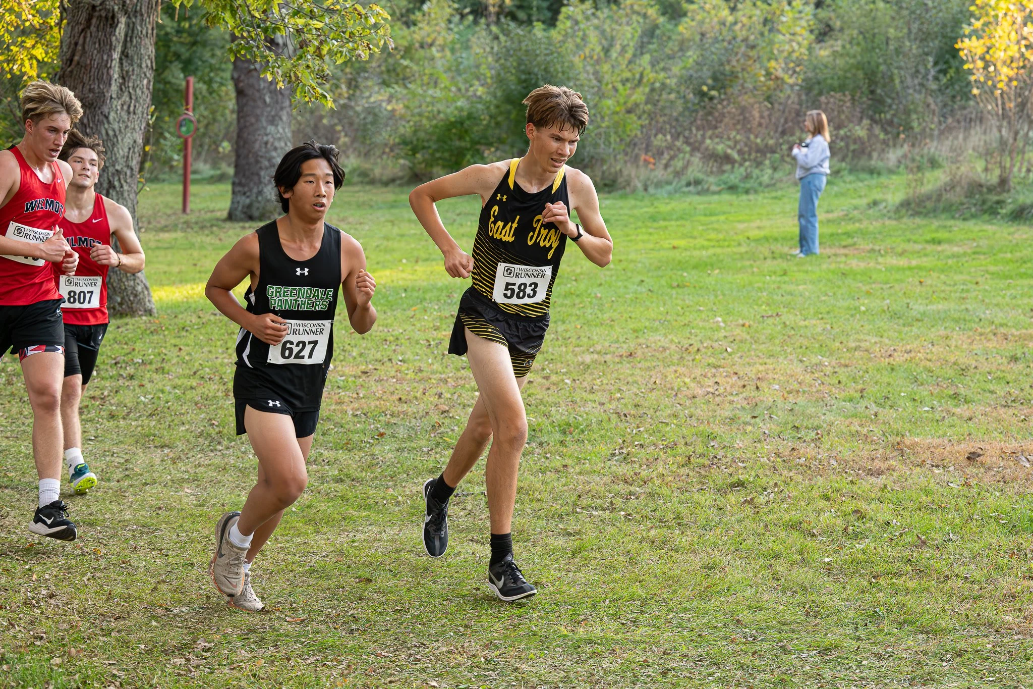 Several young male runners competing in a cross-country race on a grassy trail in a wooded area, with one runner in front and others following behind, and with a woman taking photos in the background.