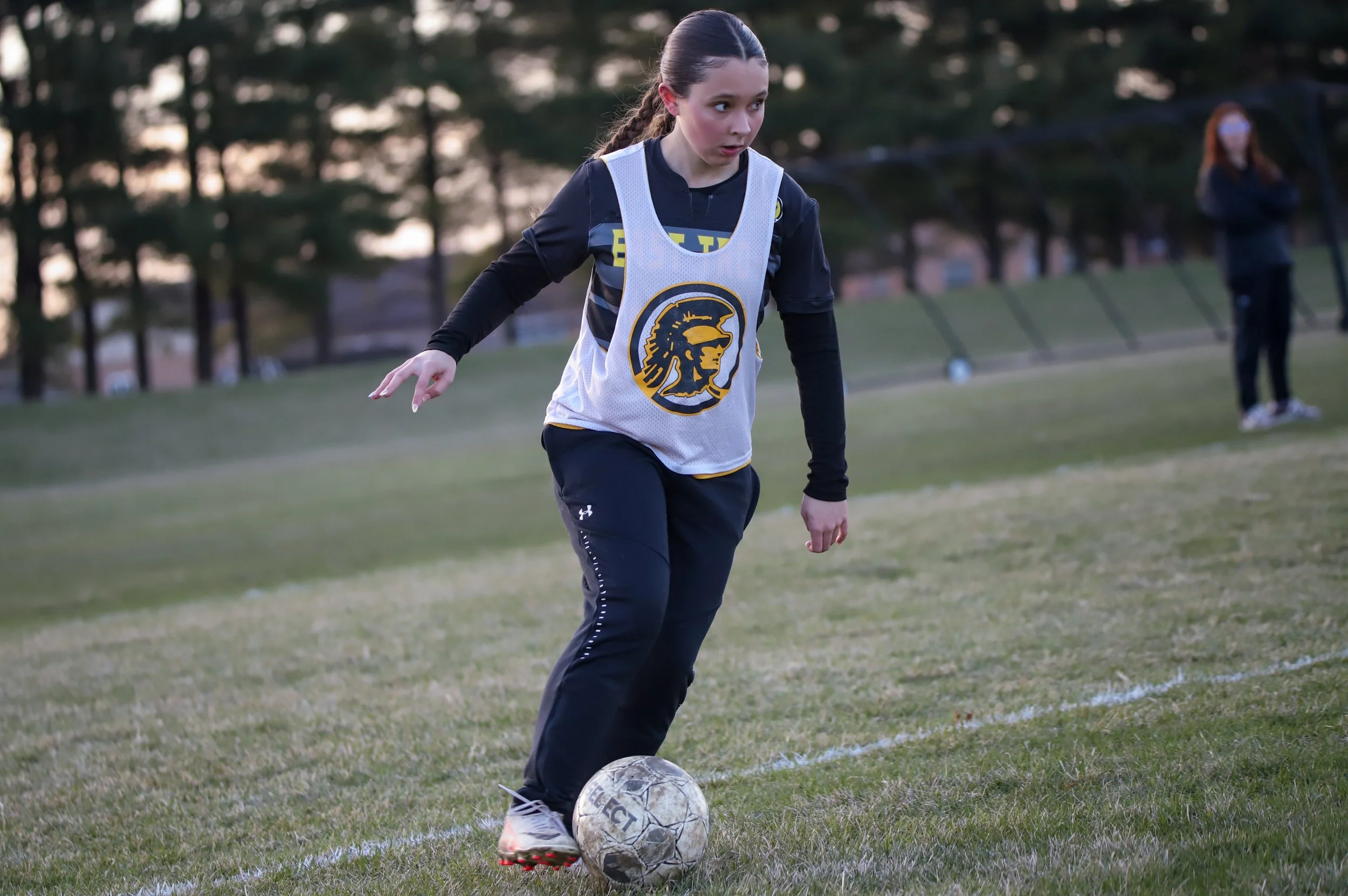 A young female soccer player with brown hair tied back, wearing a white training bib with a logo of a warrior helmet, black long-sleeve shirt, and black pants, is preparing to kick a worn soccer ball on a grassy field during late afternoon or early evening.