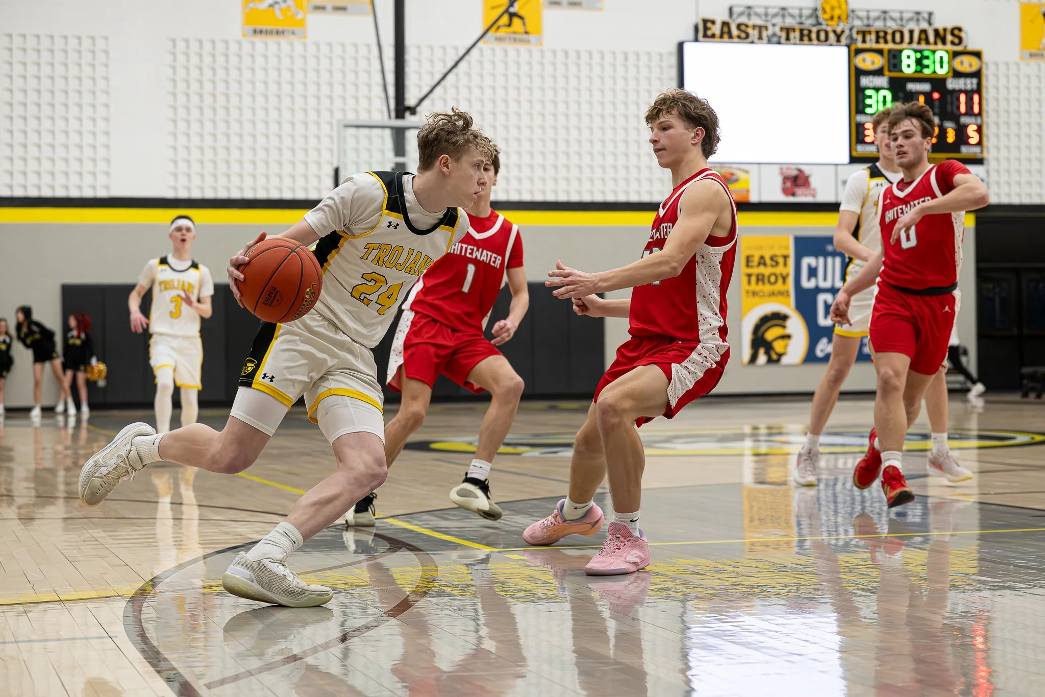 A basketball game between East Troy Trojans in white uniforms and Whitewater in red uniforms, with players in action on the court and a scoreboard showing 8:30 remaining.
