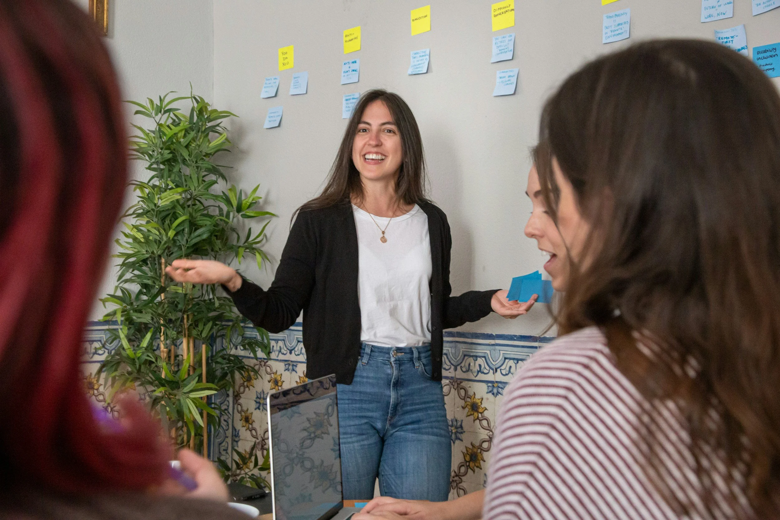 A woman leading a discussion in a meeting room with sticky notes on the wall, surrounded by seated women and a laptop.