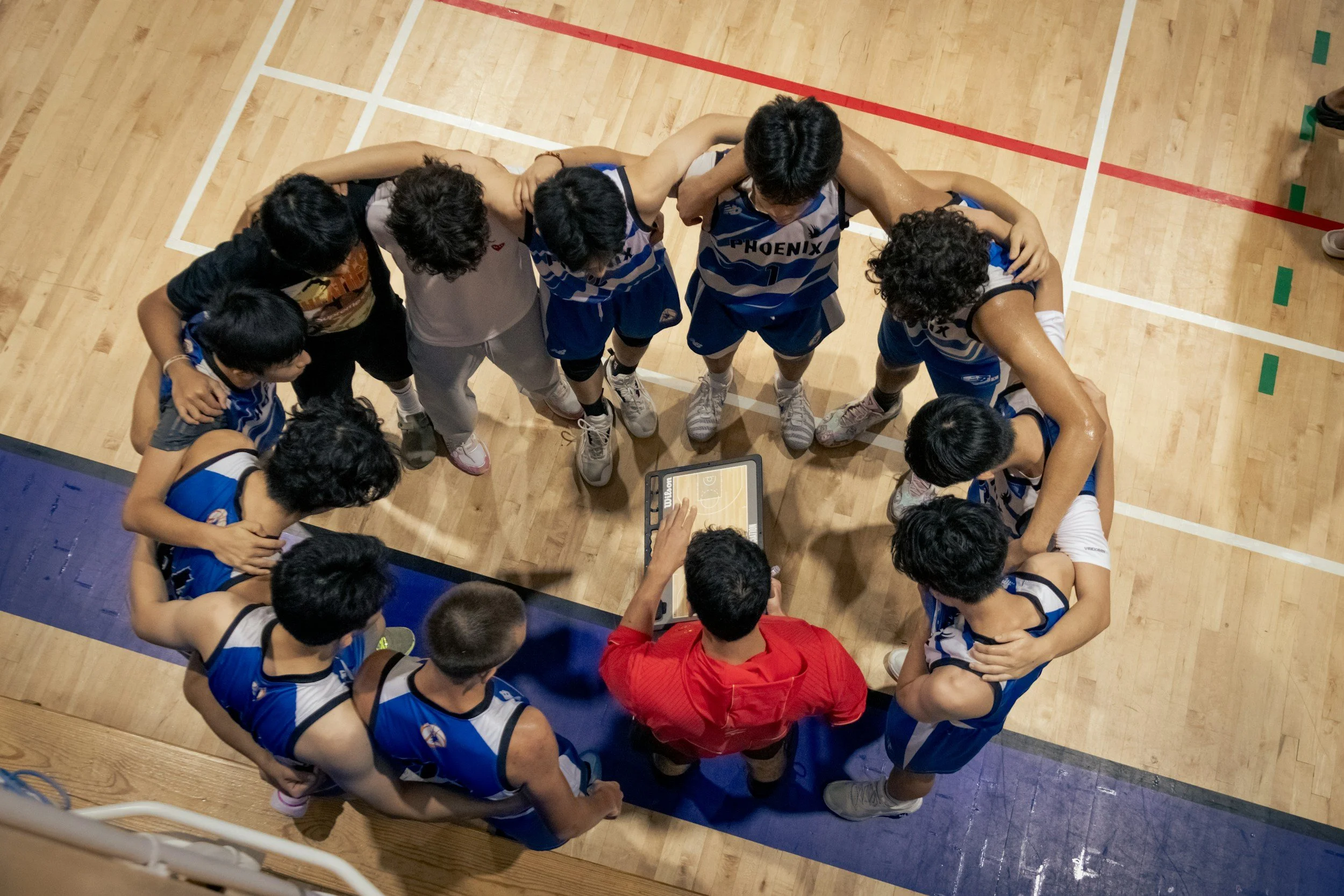 A team of basketball players in blue and white uniforms gathered around a coach in a red shirt on a basketball court, listening during a timeout.