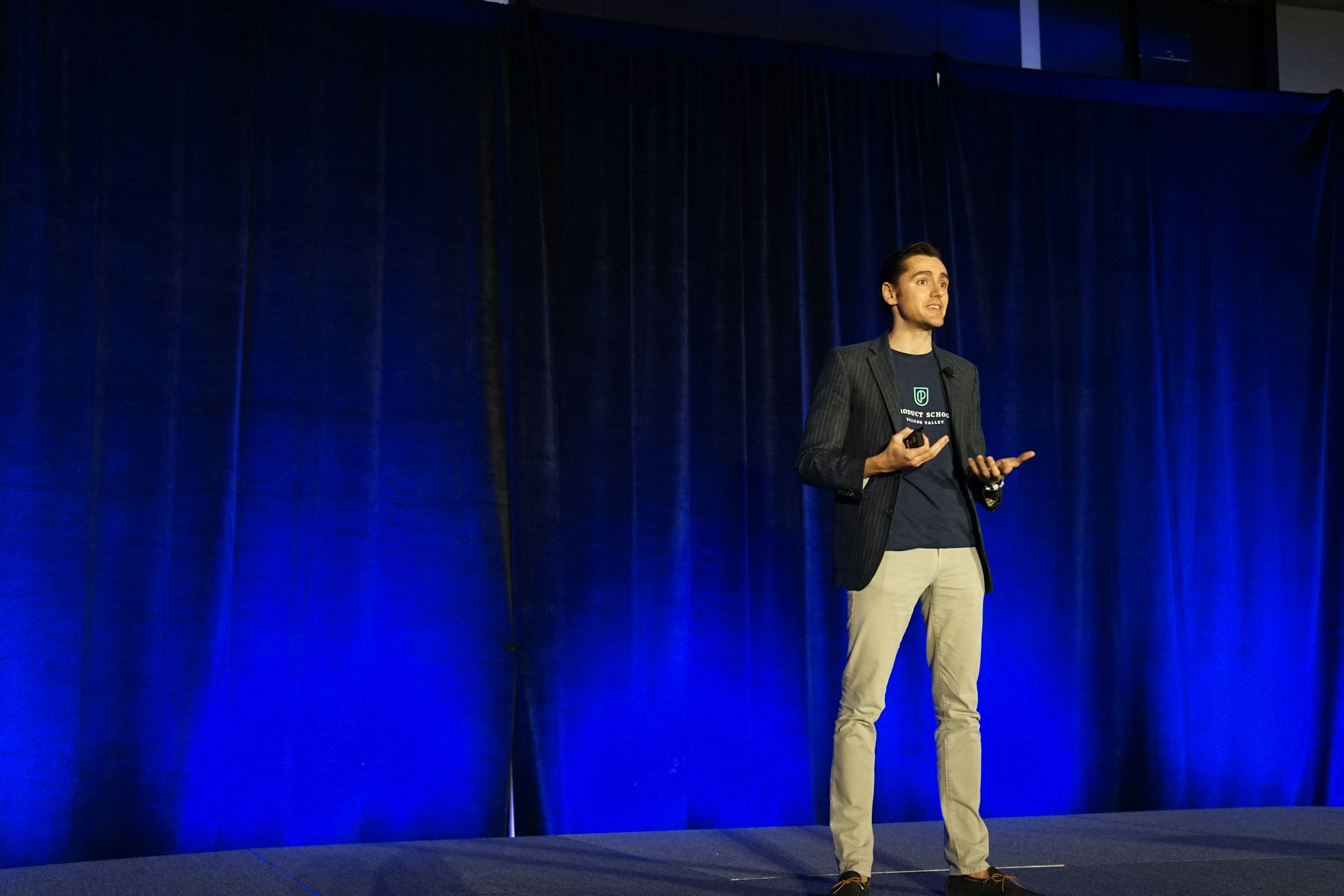 A man in a blazer and khakis gives a presentation on a stage with blue curtains background.