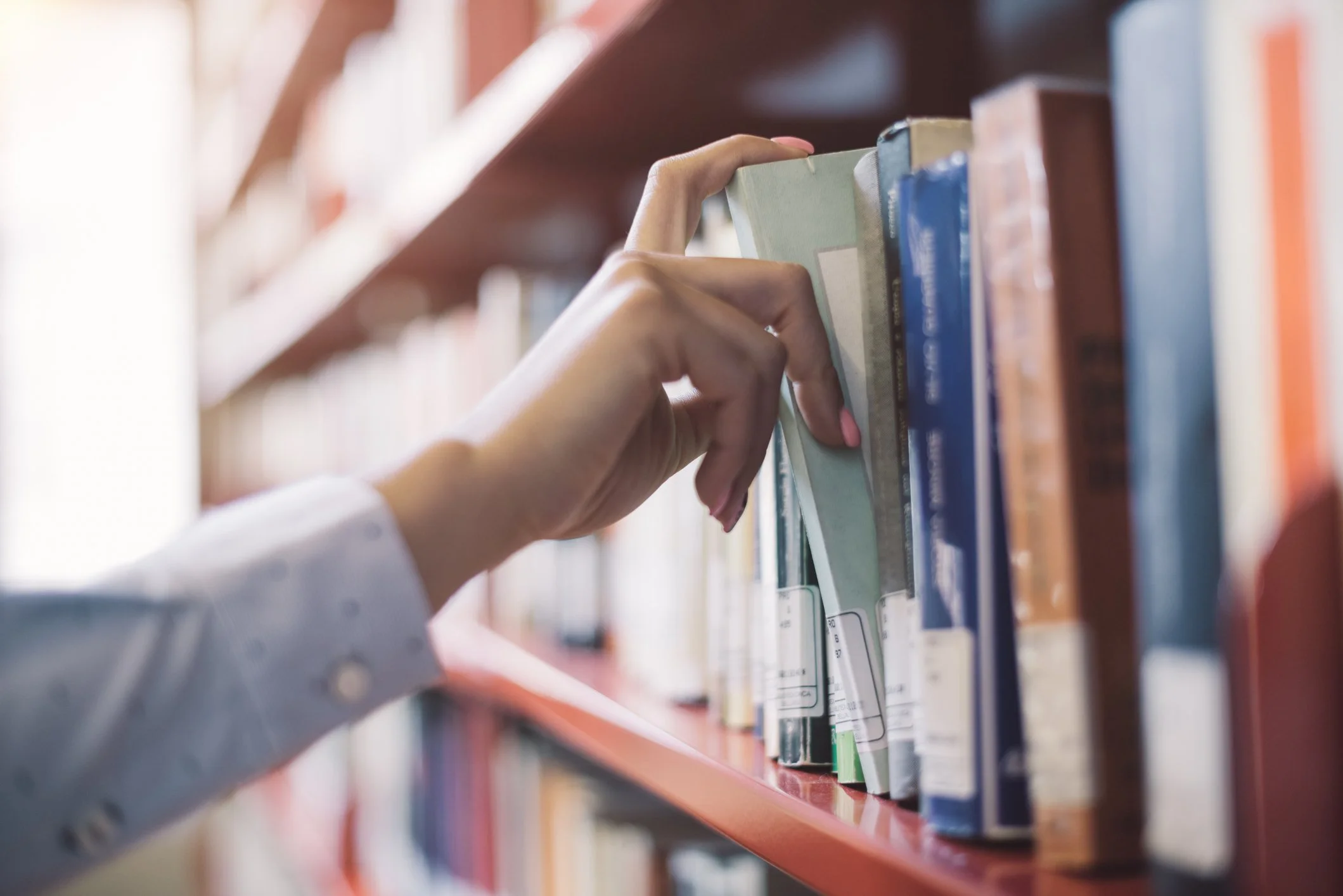 A person with light pink painted nails and wearing a white shirt is reaching for a book on a library shelf.