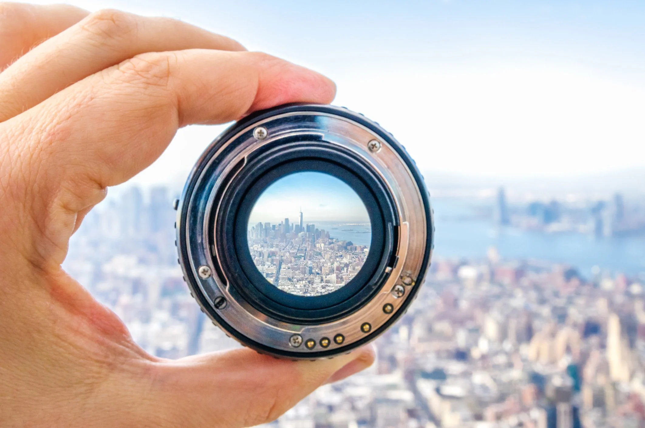 A hand holding a camera lens with a city skyline and river visible through the lens, showing a clear view of a tall building in the distance.