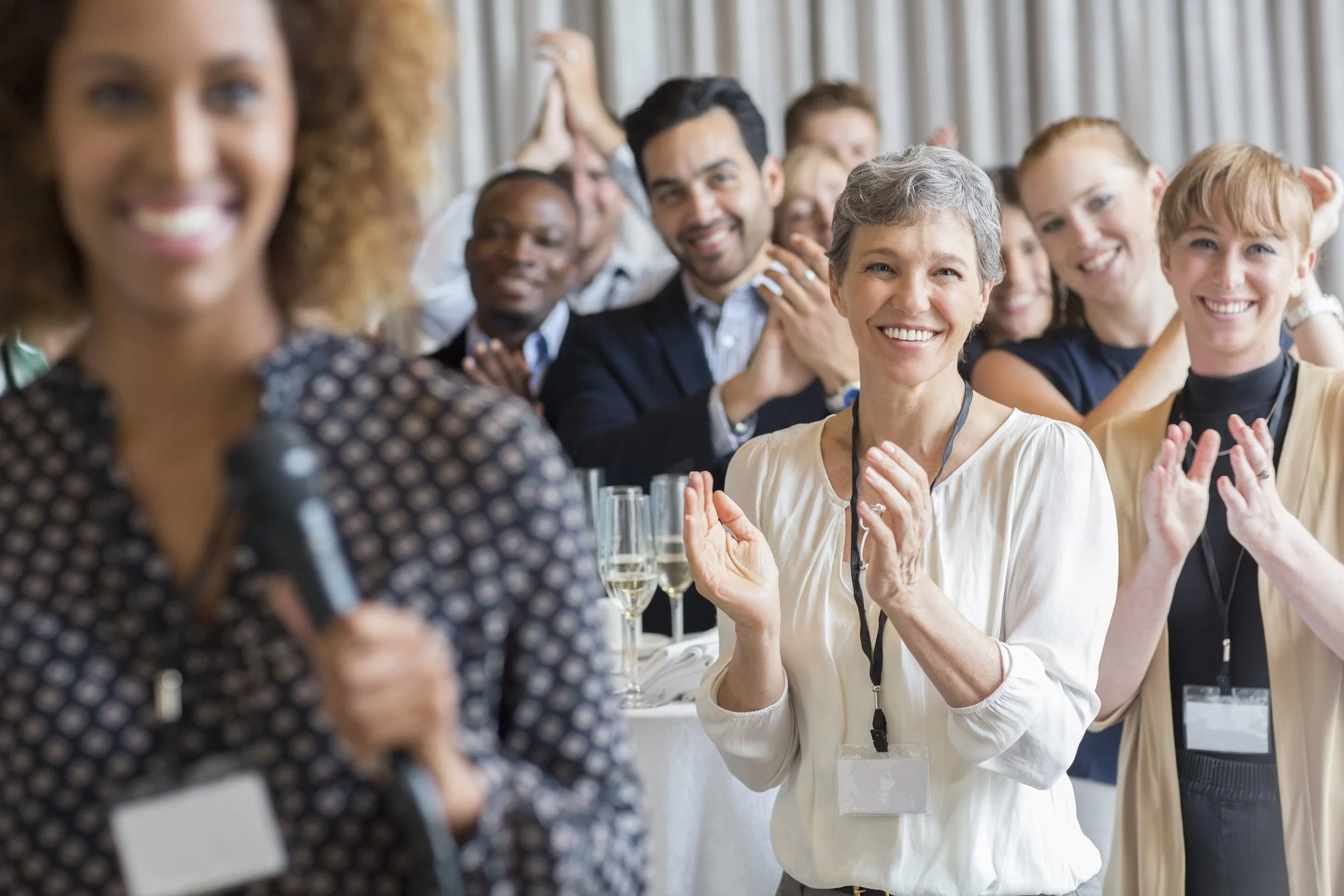 Group of diverse people at a formal event, smiling, clapping, and listening to a woman speaking into a microphone