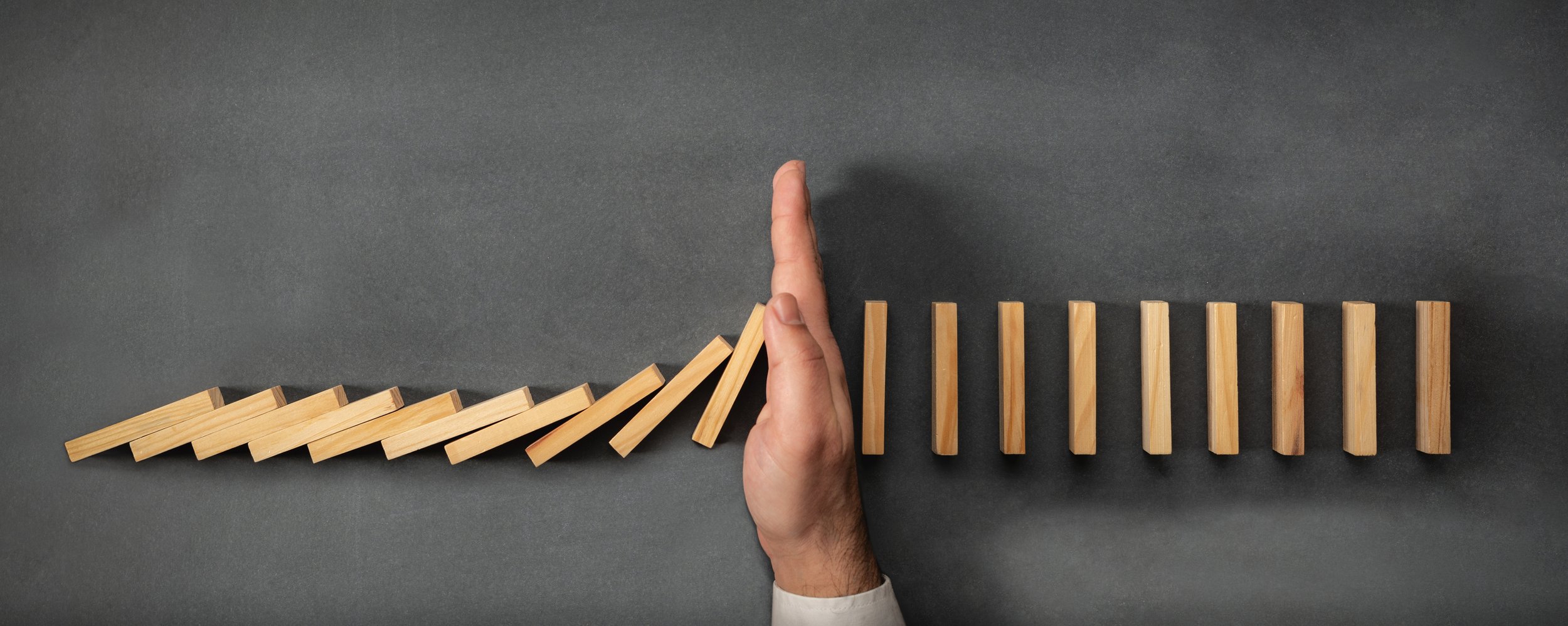 A hand stops a row of falling wooden blocks from knocking over a row of upright wooden blocks on a dark gray surface.