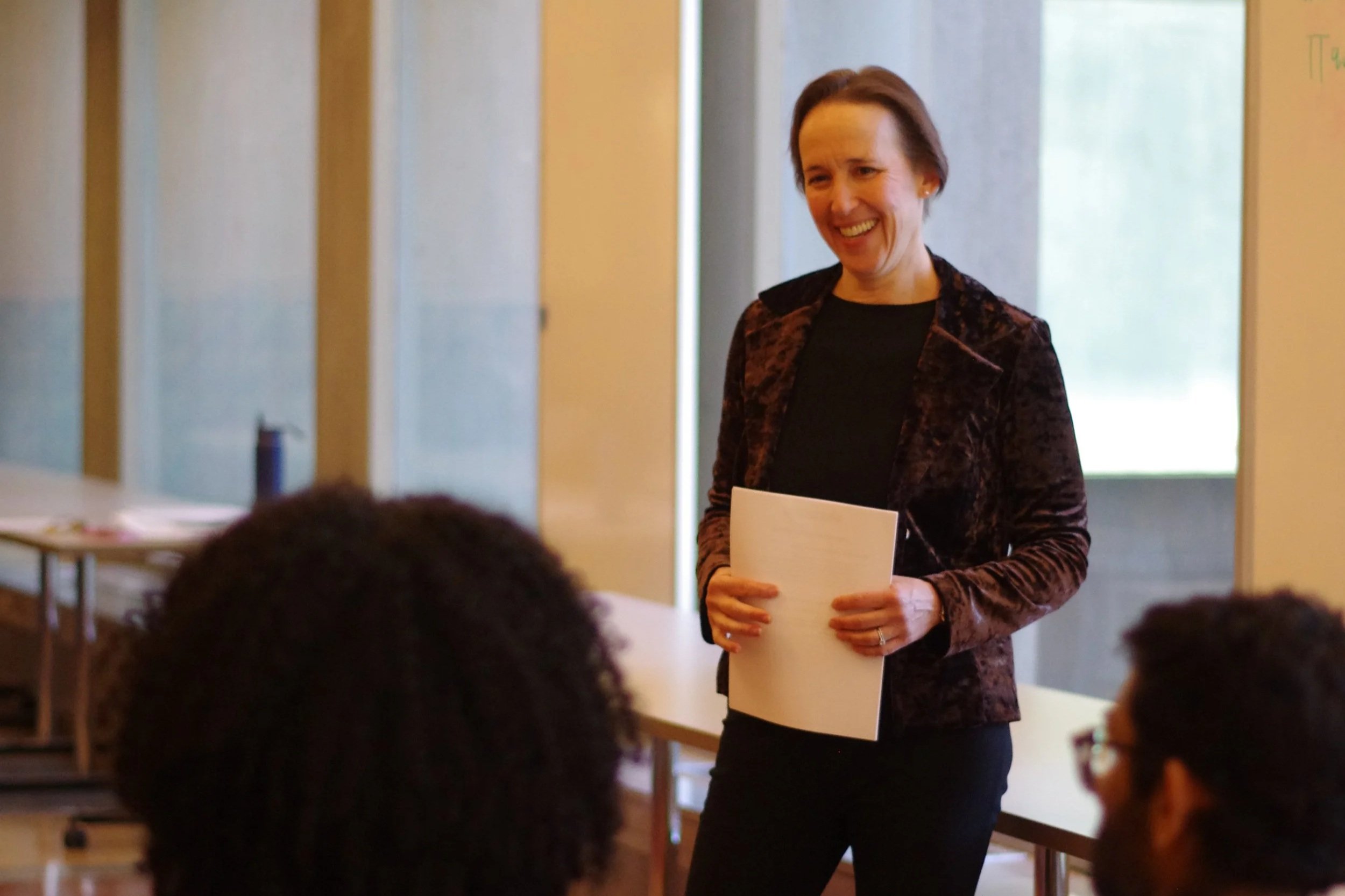 A woman with short brown hair, wearing a black shirt and a brown velvet blazer, holding papers, smiling and speaking to a group in a classroom or seminar room with large windows.