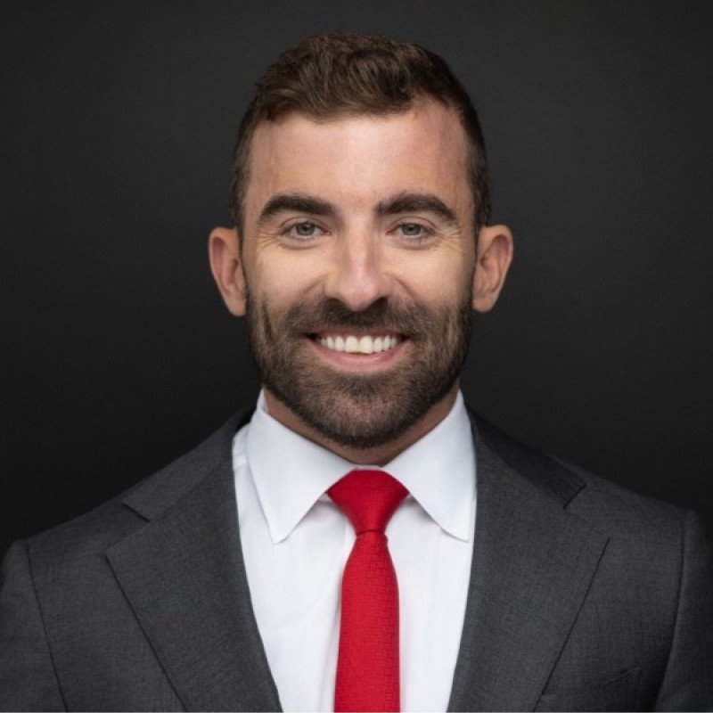 A smiling man with brown hair and a beard, wearing a dark gray suit, white shirt, and red tie, against a black background.