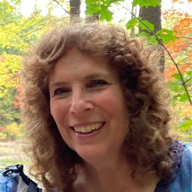 A woman with curly, shoulder-length brown hair smiling outdoors in a wooded area with fall foliage.