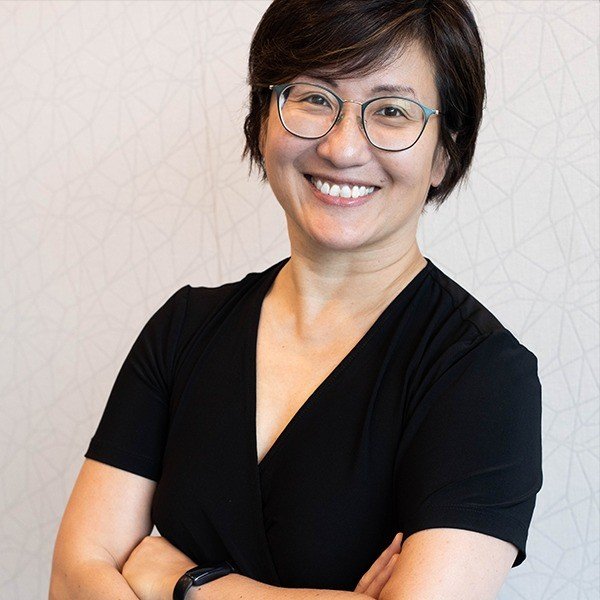 Smiling woman with short dark hair, glasses, and a black top, arms crossed, standing in front of a light patterned wall.