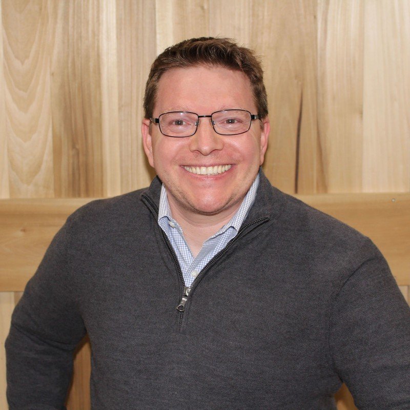 A smiling man with glasses wearing a dark zip-up sweater over a collared shirt, standing in front of a wooden wall.