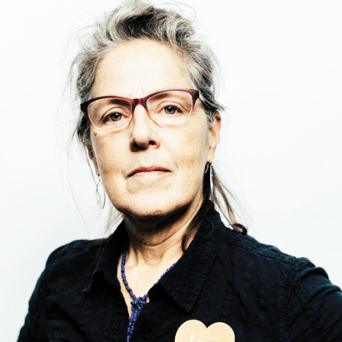 An older woman with gray hair, wearing glasses and a dark shirt, looking confidently at the camera.