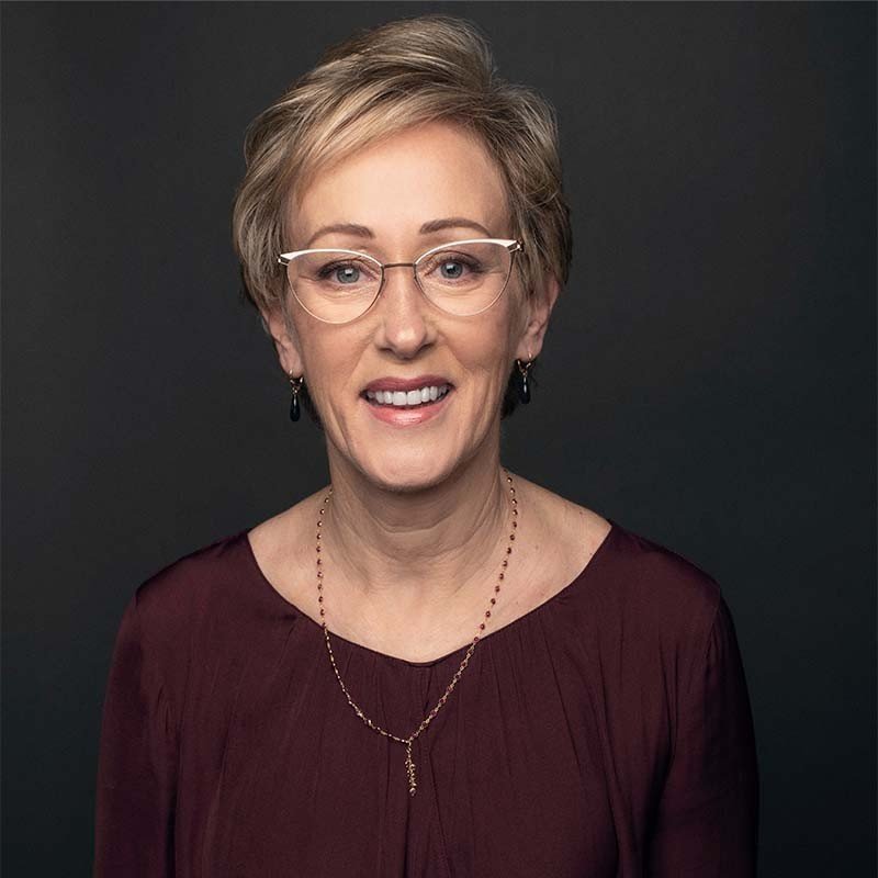 A smiling woman with short blonde hair, glasses, earrings, a maroon top, and a necklace, standing against a dark background.