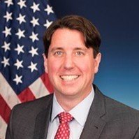 A man in a suit smiling in front of an American flag.