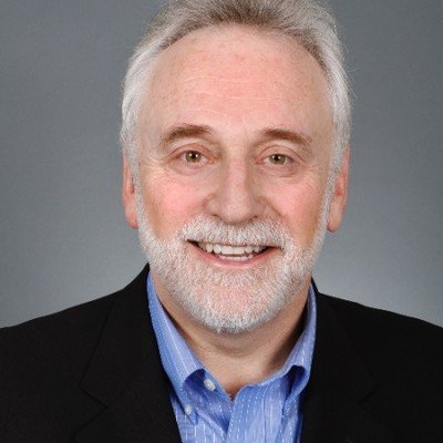 Headshot of an older man with gray hair and a beard, wearing a black blazer and blue shirt, smiling against a gray background.