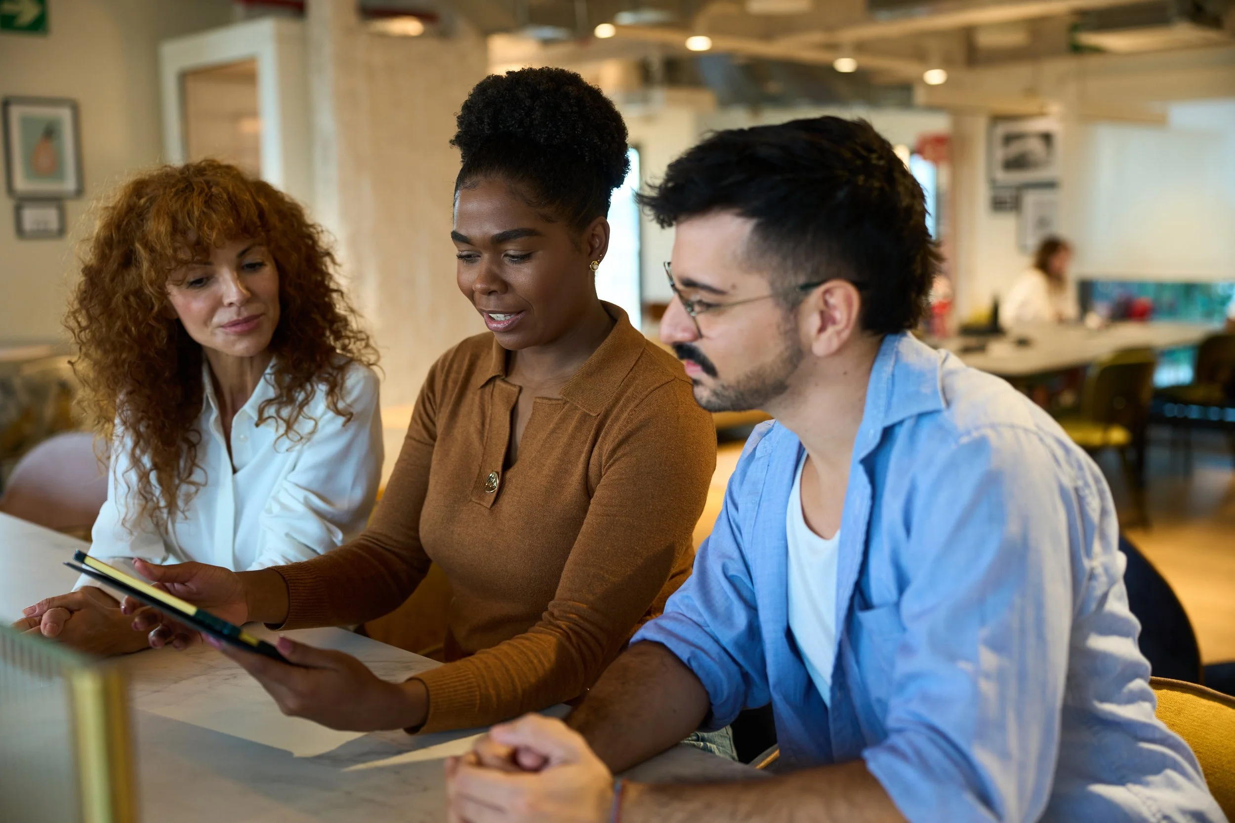 Three people sit at a table in a cafe, looking at a smartphone together, engaged in a discussion.
