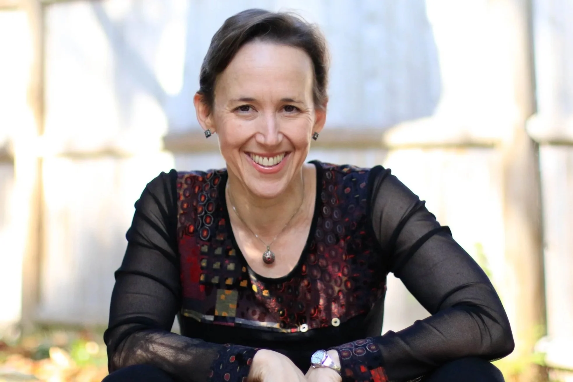 Smiling woman with short dark hair wearing a black and colorful patterned top, jewelry, and sitting outdoors.