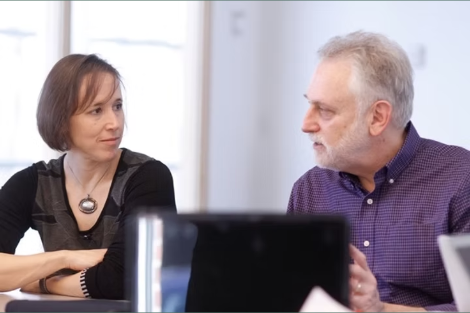 Two adults, a woman and a man, are sitting at a table engaged in a conversation. The woman has shoulder-length brown hair and is wearing a black top with a necklace. The man has white hair and a beard, wearing a purple checkered shirt. They are in a well-lit room with neutral background.
