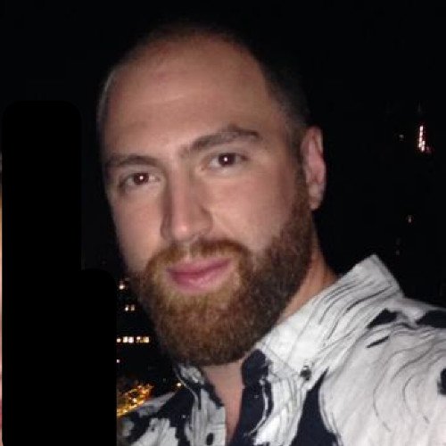 A man with a beard and short hair taking a selfie in a dark setting, wearing a patterned shirt with a cityscape in the background.