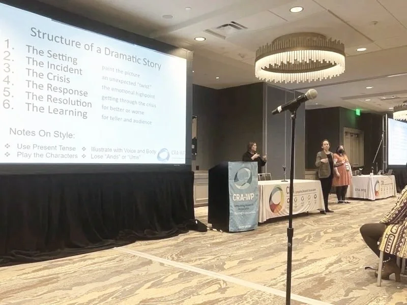 A conference room with two presentation screens, a podium, and three women standing near the tables. One woman is speaking at the podium, and a large microphone is positioned in front. The slide on the screen discusses the structure of a dramatic story, including setting, incident, crisis, response, resolution, and learning, along with style notes.
