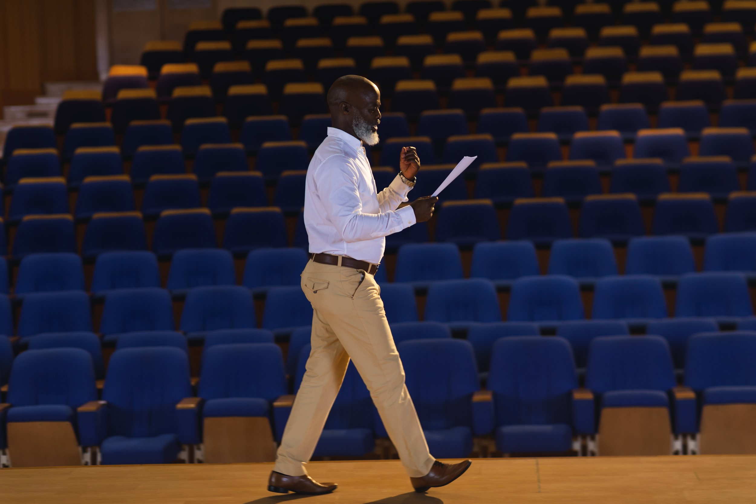 A man with a gray beard and bald head walking on a stage while reading a paper, with an empty auditorium of blue chairs in the background.