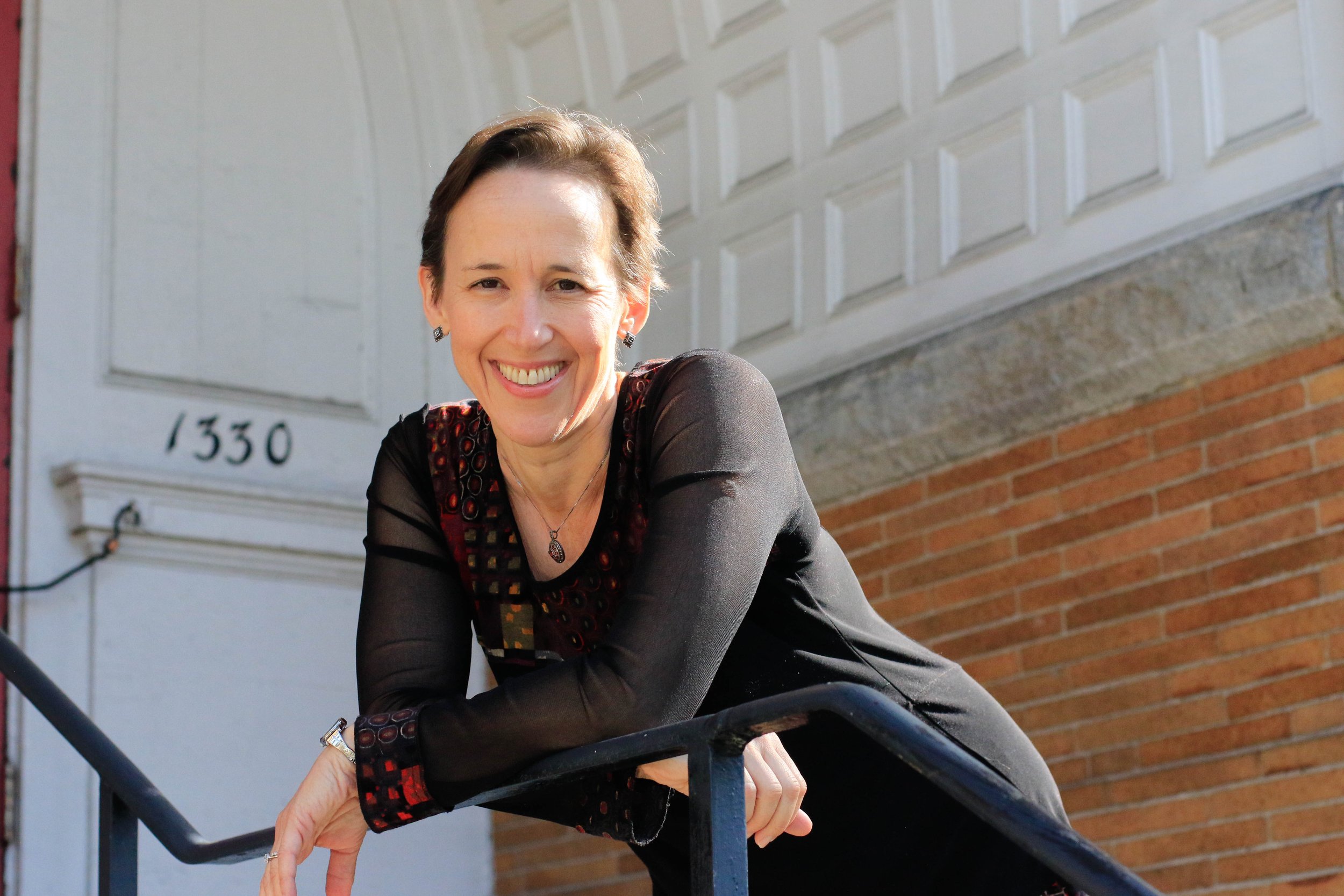 A smiling woman leaning on a black railing outside a building with the number 1330 on the wall.
