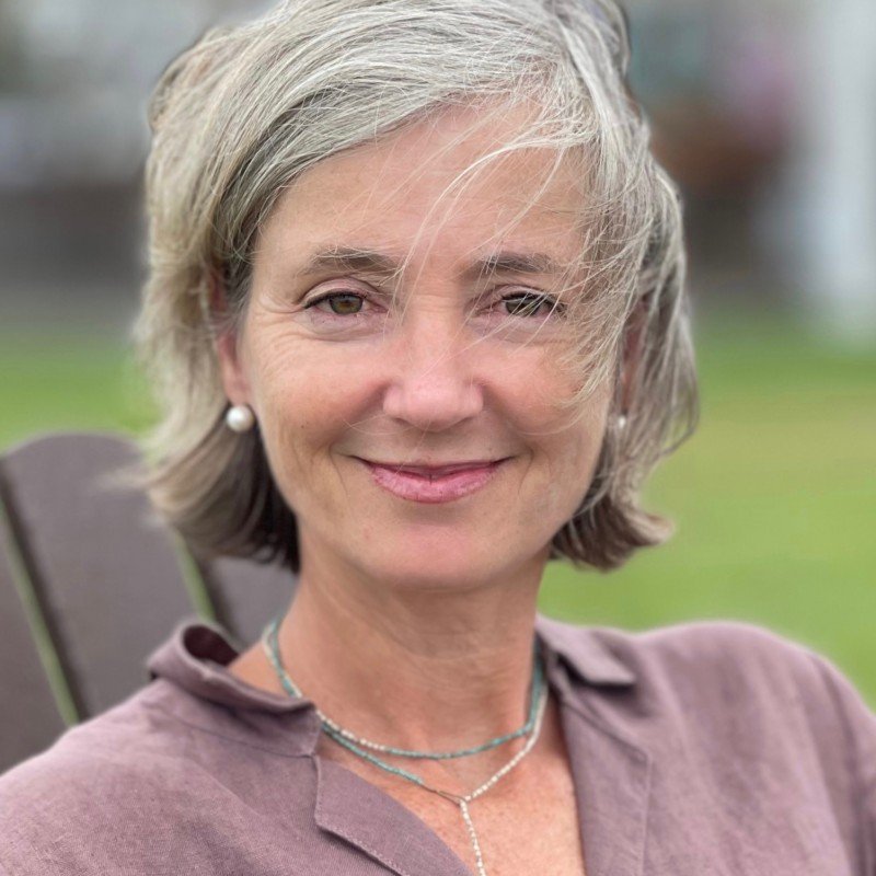 Close-up of a woman with short gray hair smiling outdoors, wearing pearl earrings and layered necklaces, with a blurred green background.