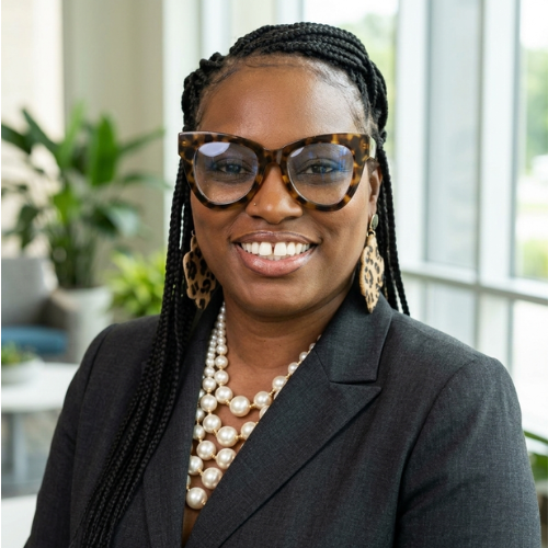 Woman in glasses smiling indoors with large windows and green plants behind her.