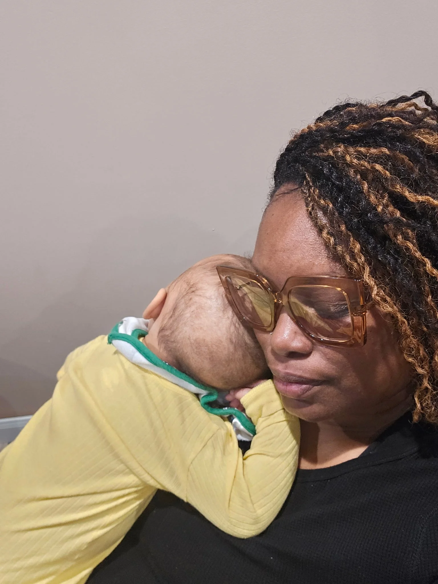 A woman wearing large brown glasses with textured hair holding a small child with light skin, wearing a yellow outfit with green and white collar, resting her head on her shoulder.