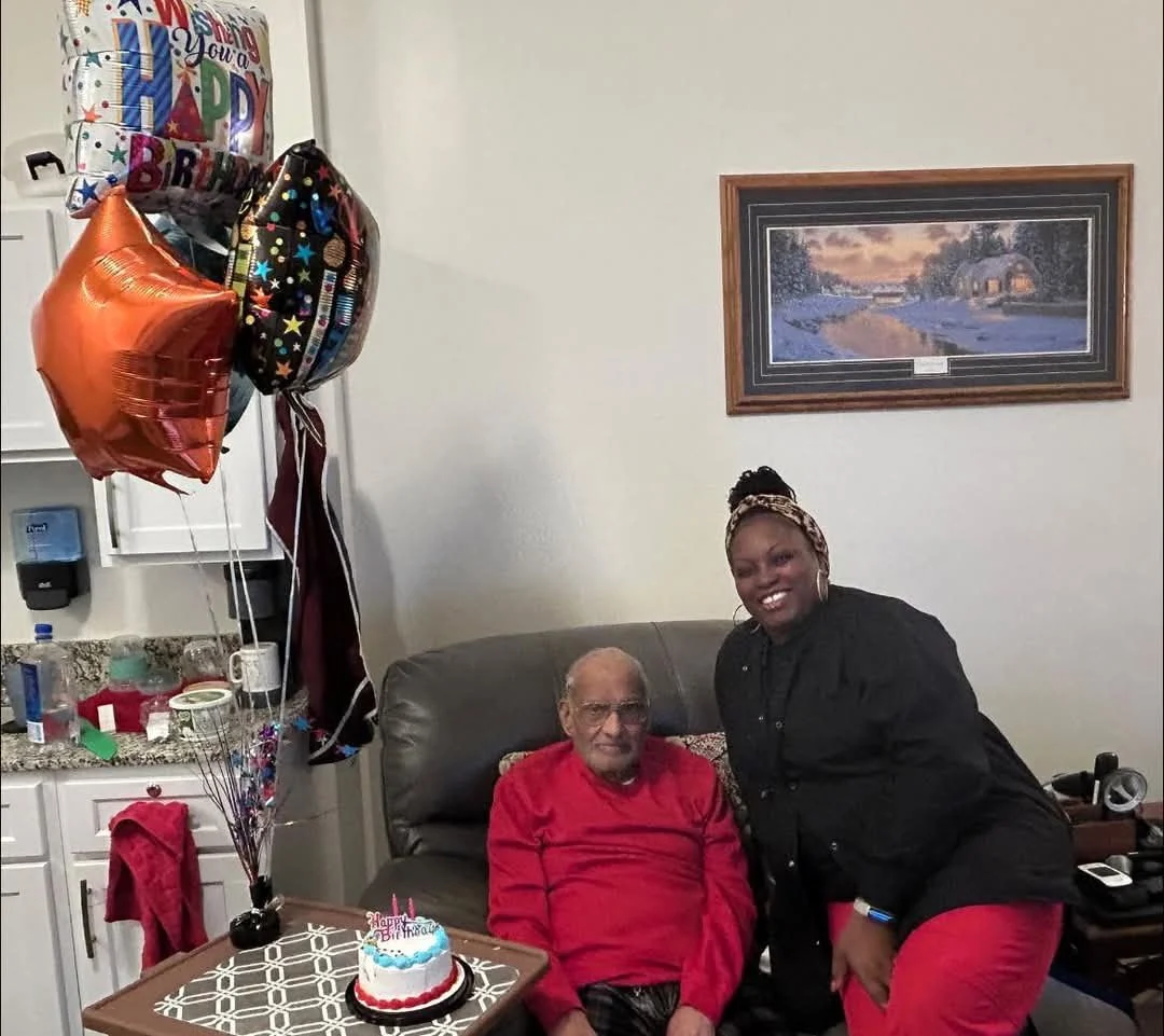 A birthday celebration with a man sitting on a sofa and a woman standing next to him. There are balloons including one with a 'Happy Birthday' message, and a small birthday cake with candles on a table.