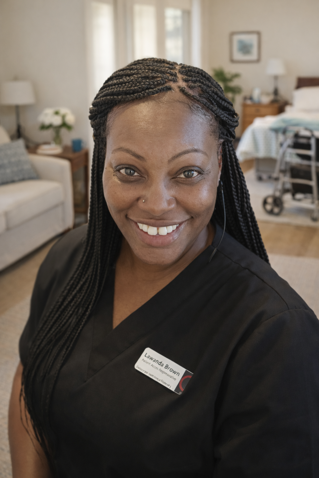 A woman with braided hair smiling at the camera, wearing a black uniform with a name tag that reads Lawanda Brown, in a cozy living room with a beige couch, lamp, and a bed in the background.
