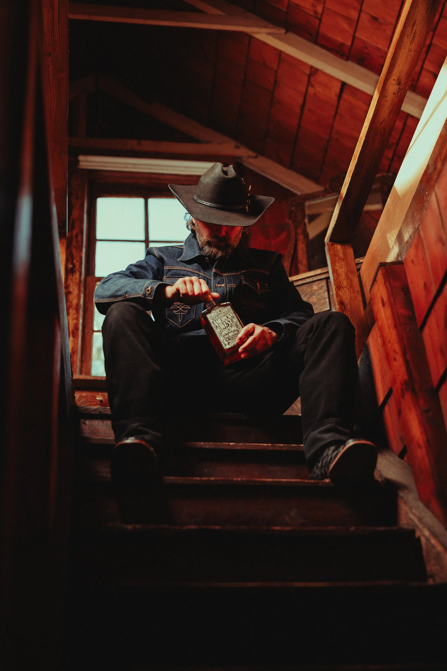 A man wearing a black cowboy hat, denim jacket, and black pants sitting on wooden stairs inside a rustic cabin. He is opening a small red box, with a window in the background letting in natural light.