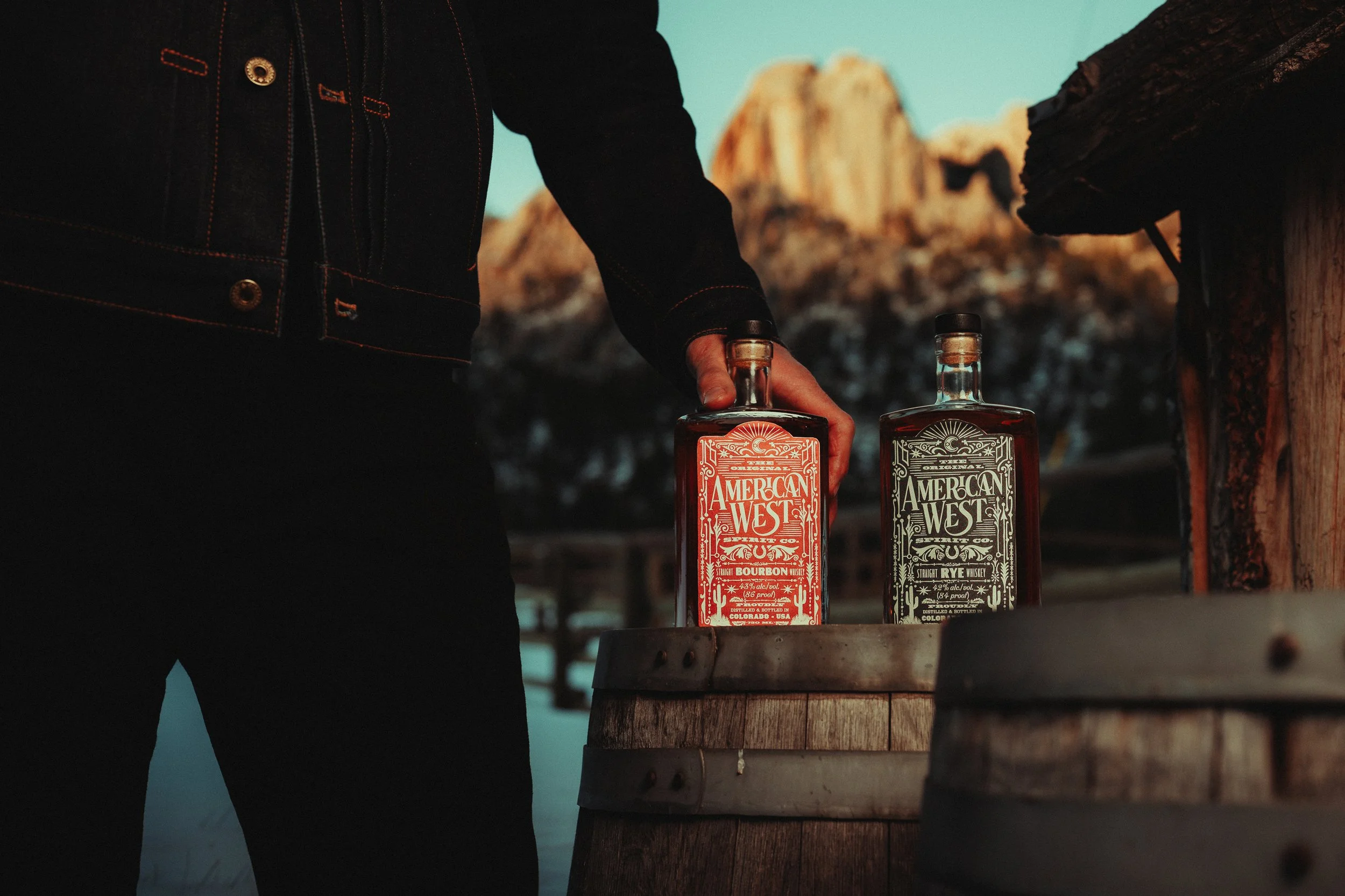 A person in a black jacket reaching for two bottles of American West whiskey placed on a wooden barrel outdoors with mountains in the background.