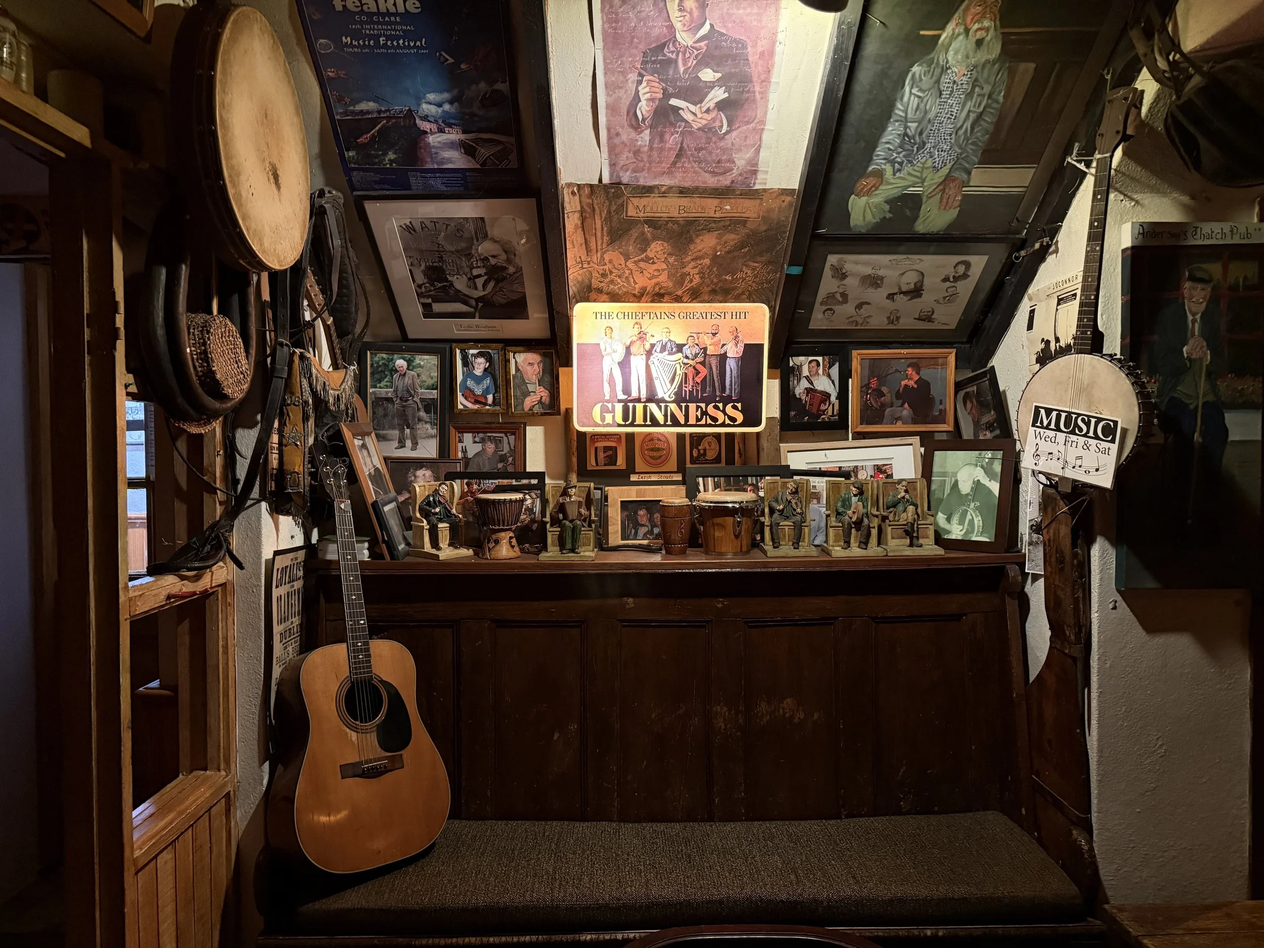 Interior of a bar or pub decorated with framed photos, posters, and musical instruments, including a guitar hanging on the left and drums on the countertop. The wall features vintage photos, a Guinness sign, and music-themed decor, creating a cozy, nostalgic atmosphere.