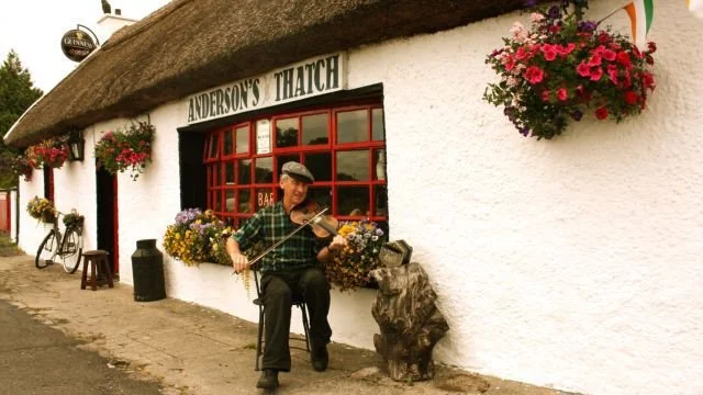 A man plays the violin outside Anderson's Thatched pub, which has a thatched roof, red window frames, and flower baskets on the white wall.