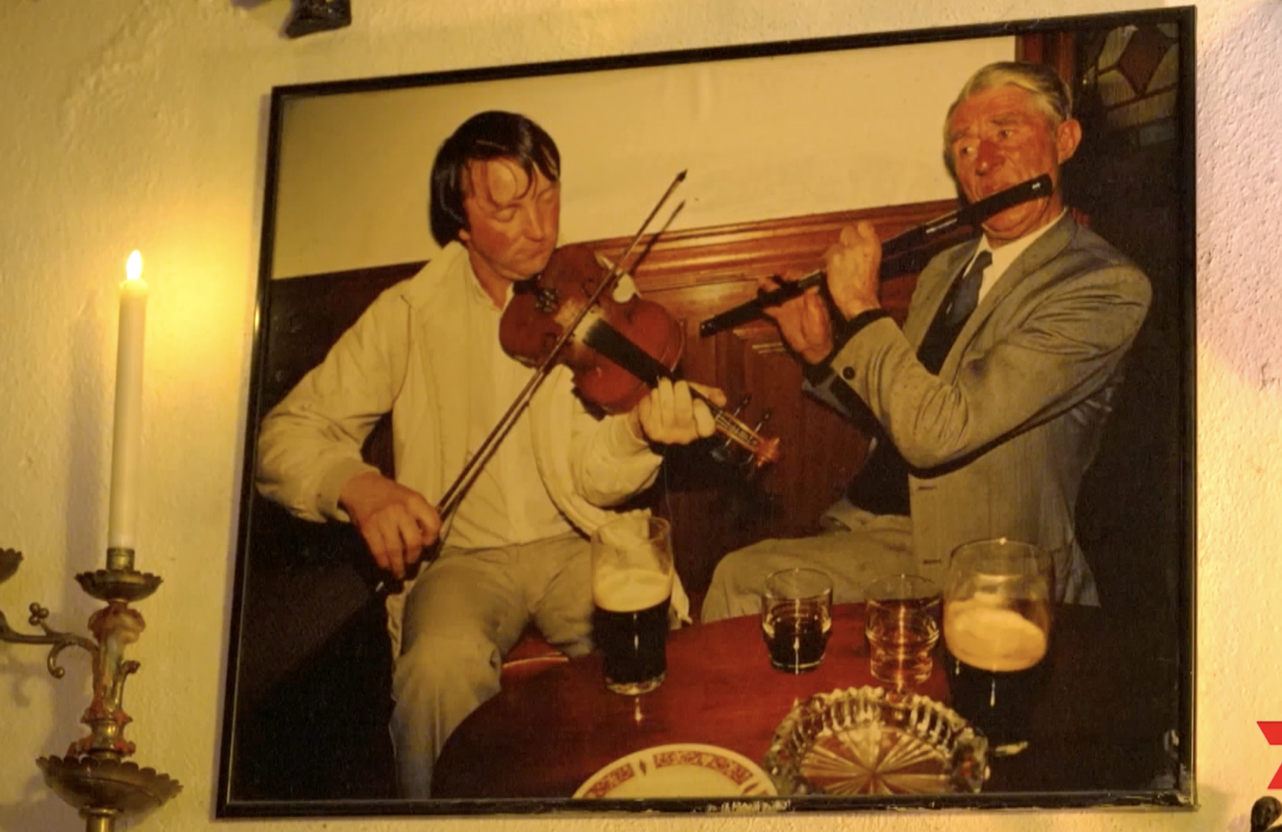 A photo of two people playing musical instruments, a woman with a violin and a man with a flute, in a cozy indoor setting with drinks on a table.