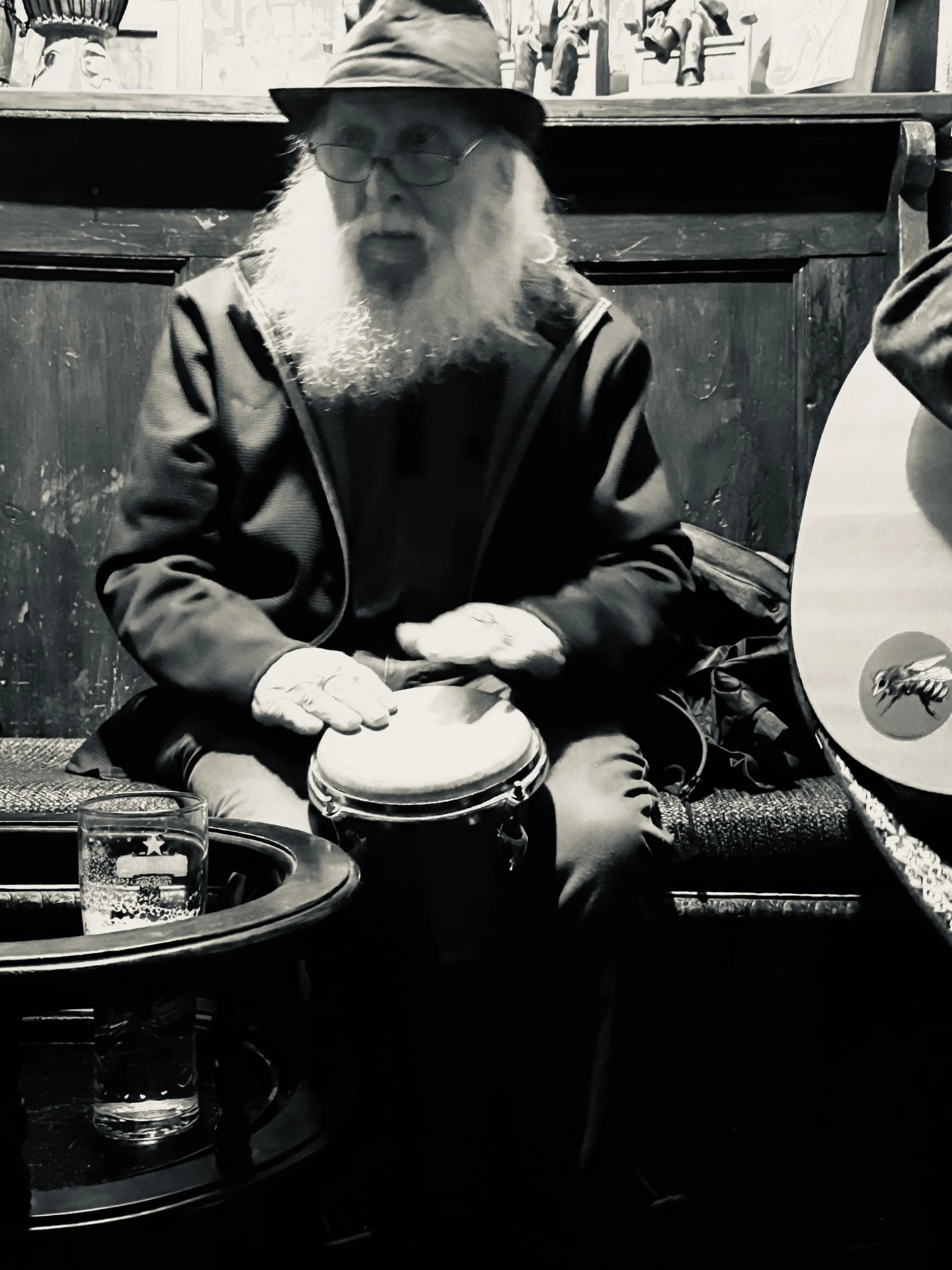 An elderly man with a long beard, glasses, and a hat, playing a hand drum while sitting in a dimly lit room. A glass of beer is on a table in front of him, and part of a guitar is visible on the right.