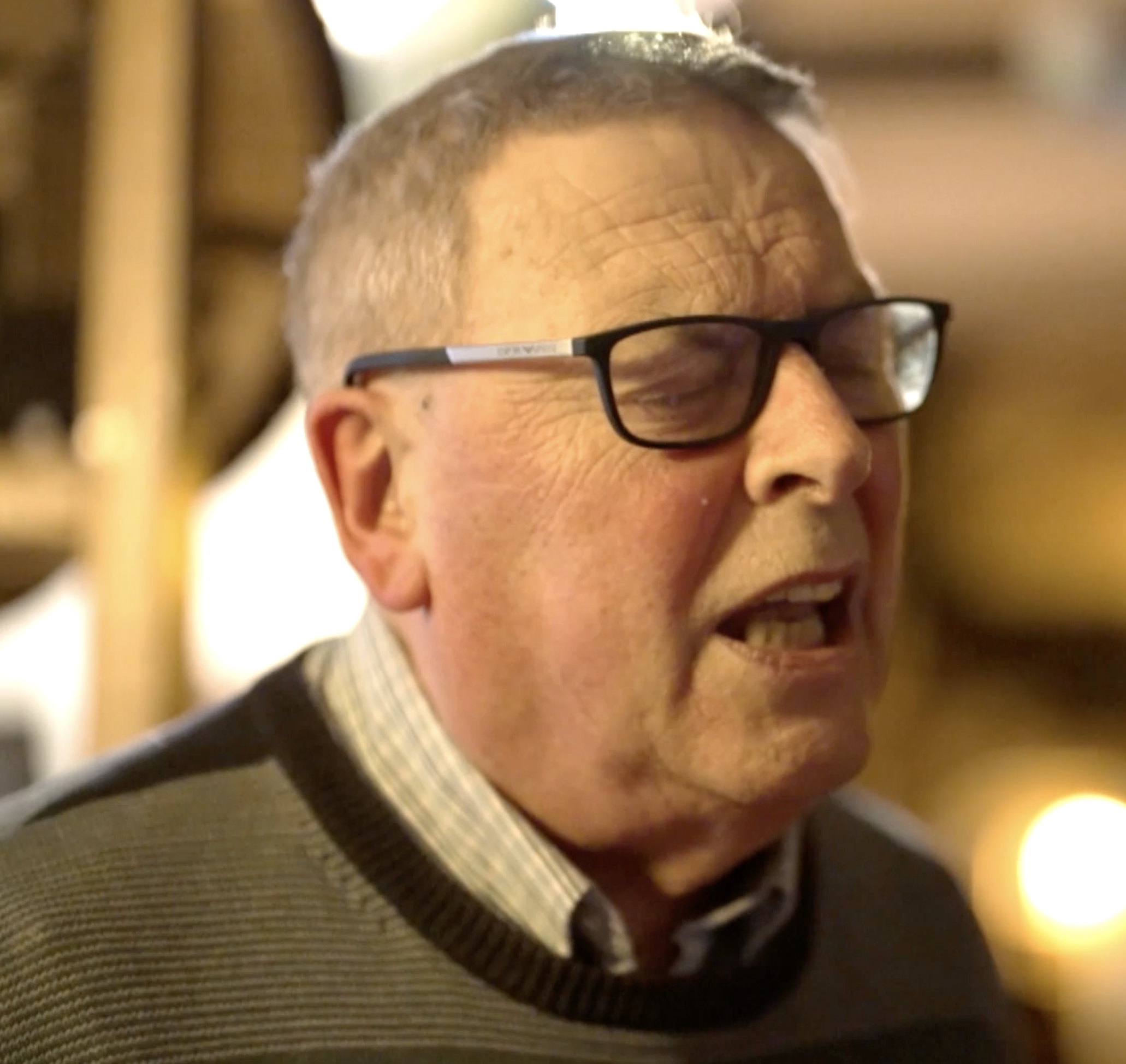 An elderly man with glasses, closed eyes, and an open mouth, possibly singing or speaking, in a warmly lit setting.